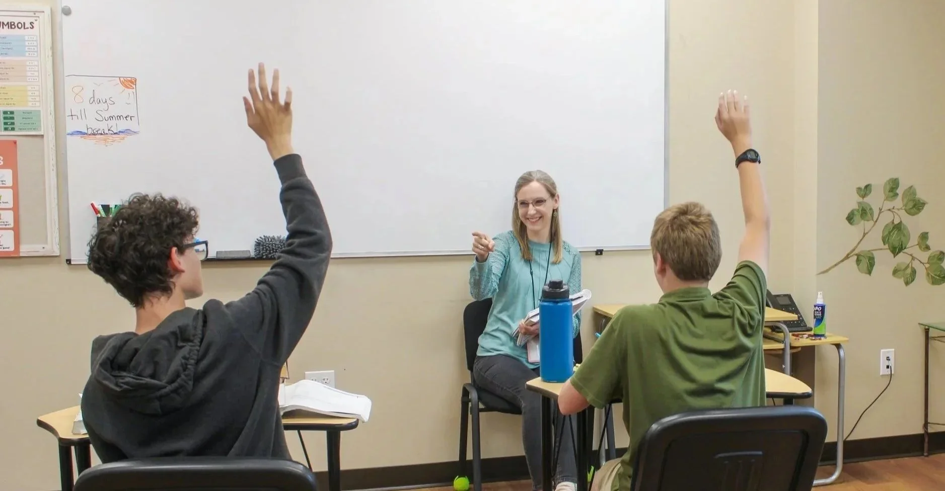 A classroom scene with a teacher and three students. Two students have their hands raised, and the teacher is smiling and pointing at one student, sitting in front of a whiteboard.