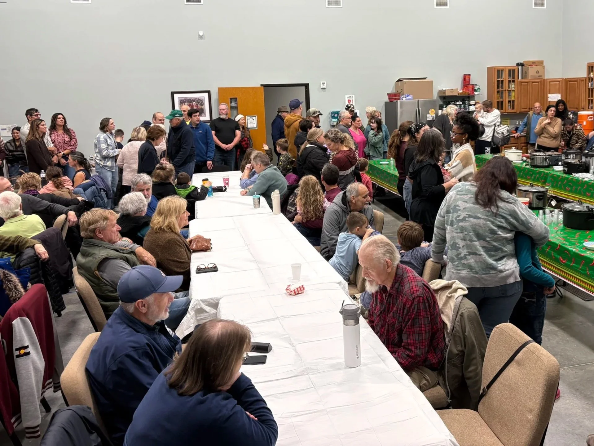 Large group of people gathered in a community center room with a serving table and kitchen area, some standing and others sitting at long tables, socializing and waiting for food or drinks.