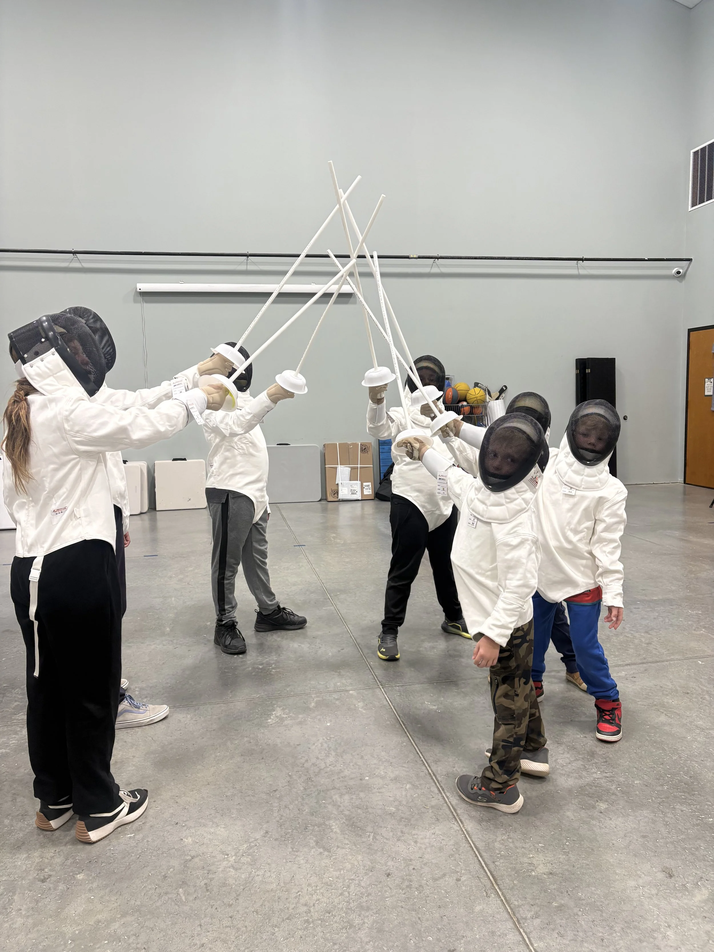 Children dressed in fencing gear practicing fencing indoors.