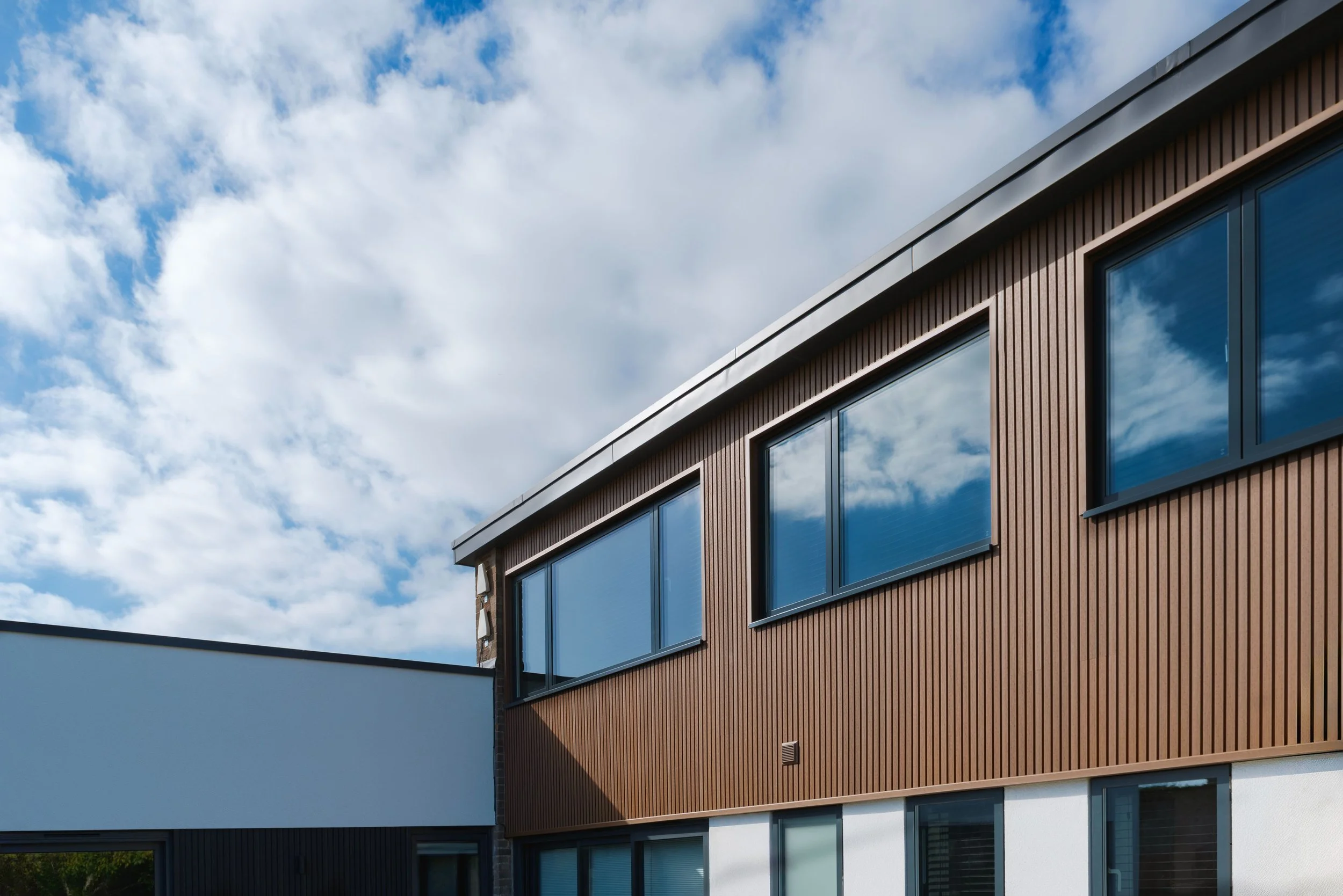 Modern multi-story building with brown and white exterior walls, large reflective windows, and a opening sky with clouds