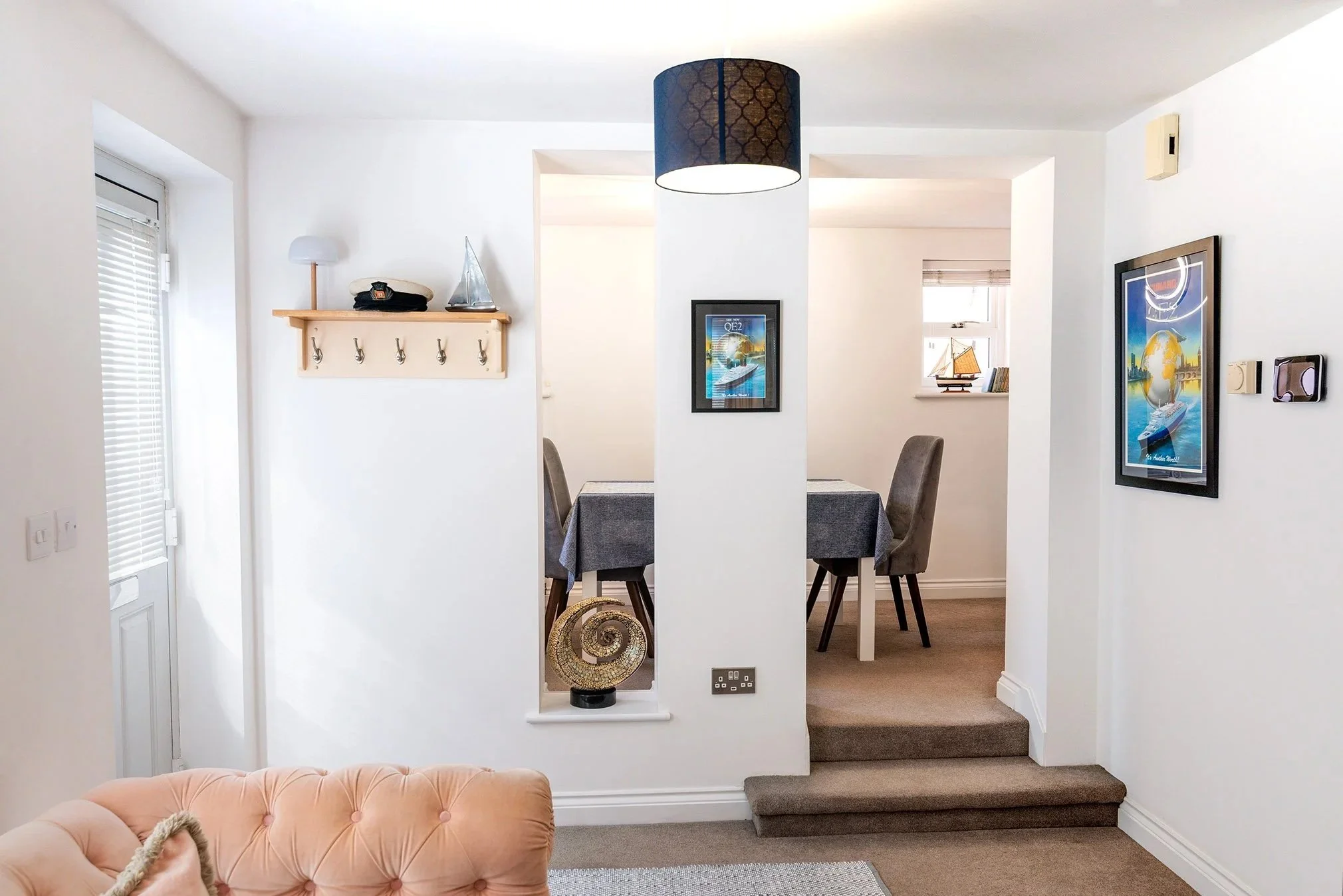 Interior view of a Brixham holiday home with white walls, a staircase with carpeted steps, framed artwork, a small window with blinds, a peach-colored tufted sofa, and decorative items including a spiraled sculpture.