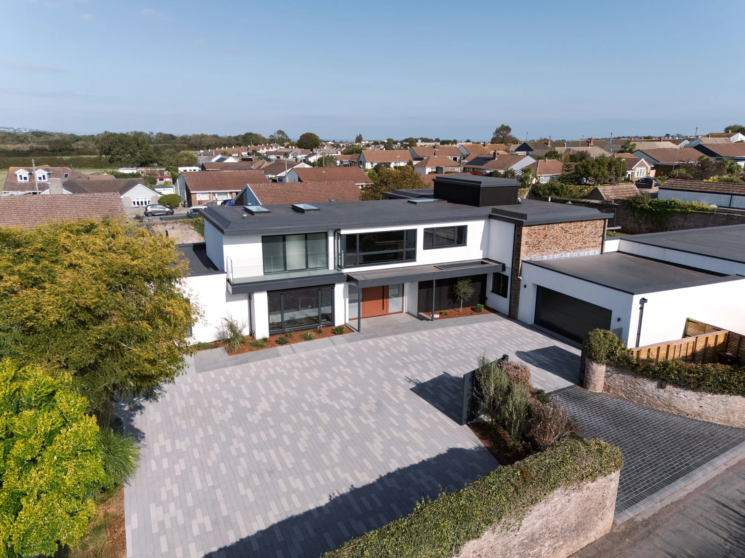 A modern two-story house with a gray roof, white walls, large windows, a wooden front door, and a spacious paved driveway, situated in a suburban neighborhood with other houses and greenery in the background.