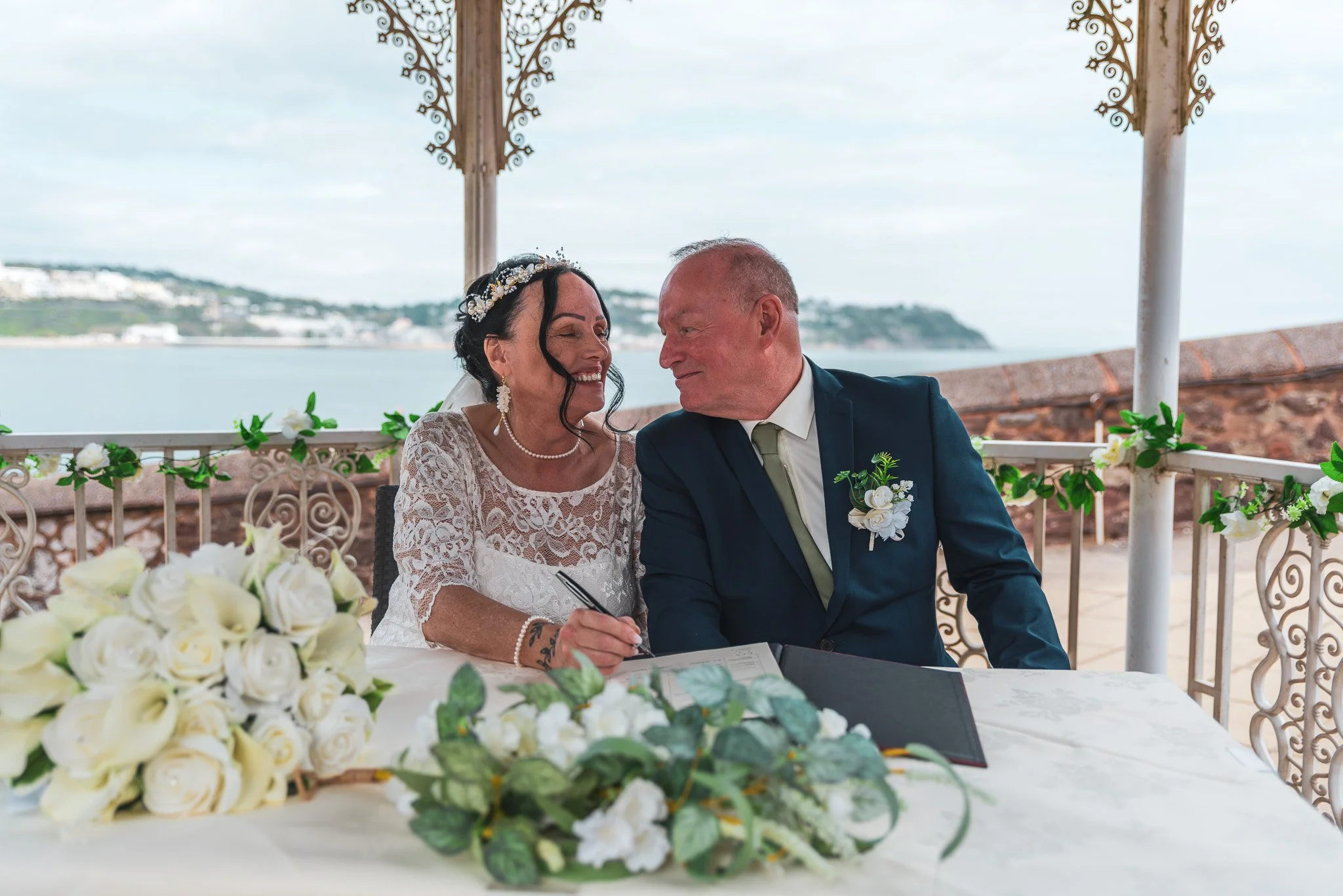 A bride and groom sit at a wedding table underneath a gazebo overlooking a body of water, with decorative ironwork and floral arrangements in the foreground.