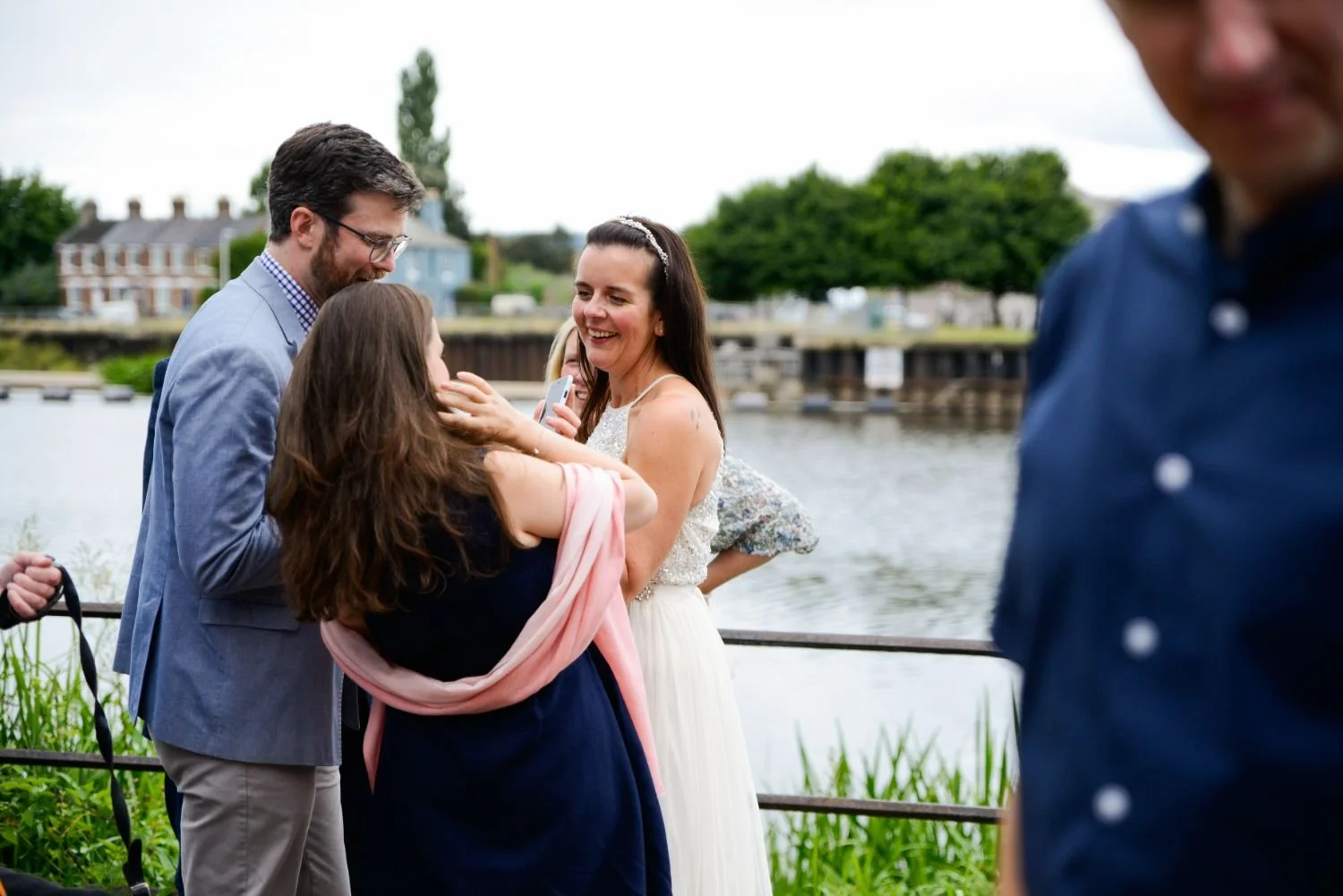 A woman in a white dress and a man in a light blue blazer are smiling and interacting with a girl near a river, with another person partially visible in the foreground.