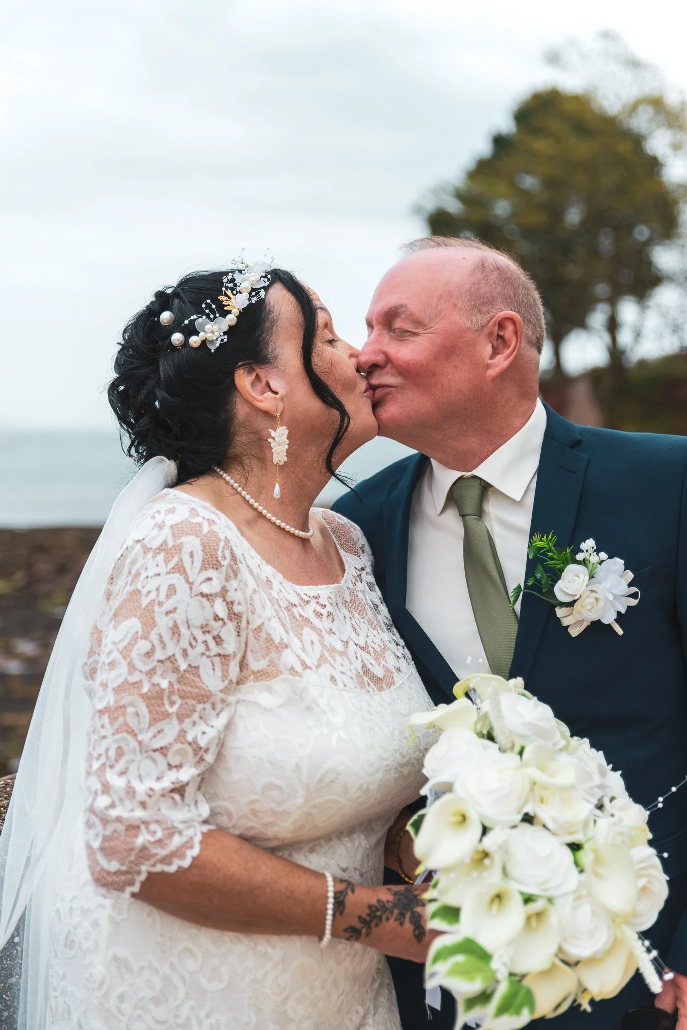 A bride and groom sharing a kiss during their wedding ceremony outdoors, with the bride holding a bouquet of white flowers.