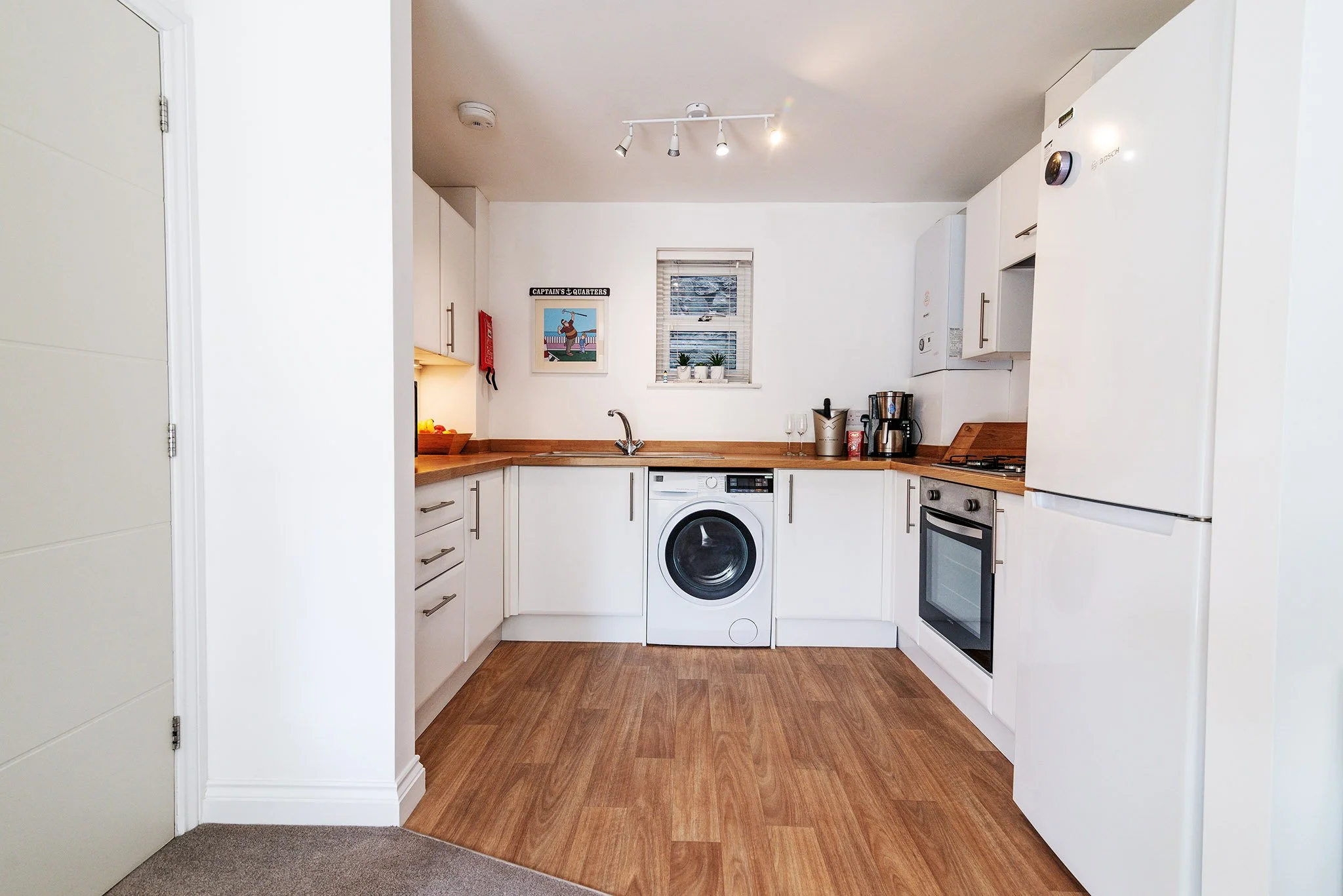 View of an open door of a modern holiday let kitchen in Brixham featuring white cabinets, a wooden countertop, a washing machine, a microwave, a coffee maker, and a window with blinds, with decorative plants and artwork.