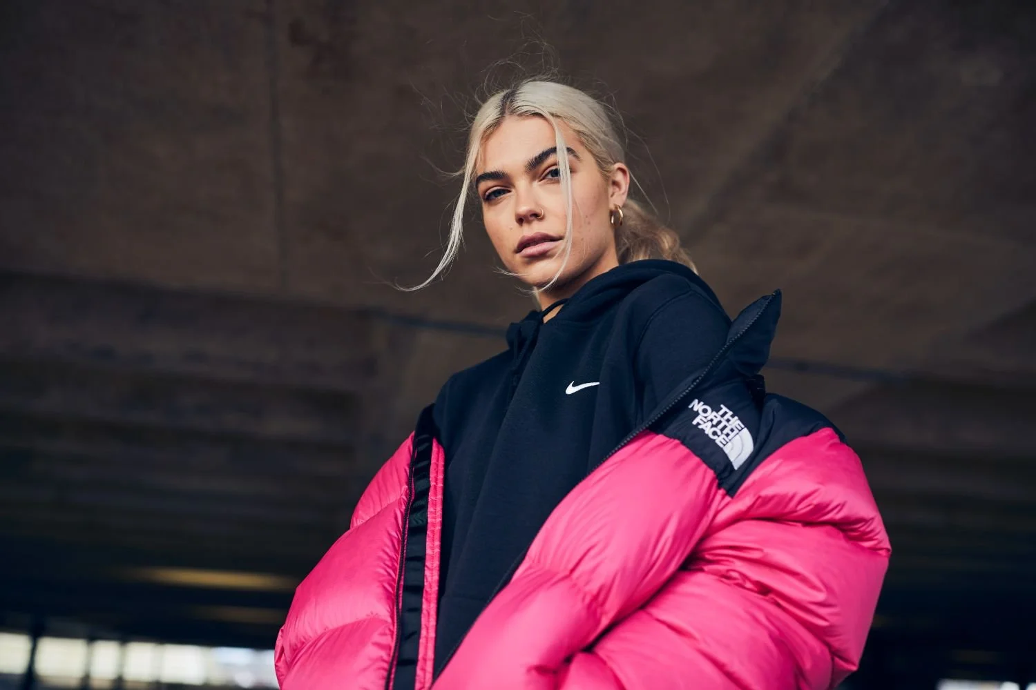 A young woman with blonde hair tucked into a hoodie, wearing a black Nike hoodie and a bright pink North Face jacket, standing under a concrete structure.