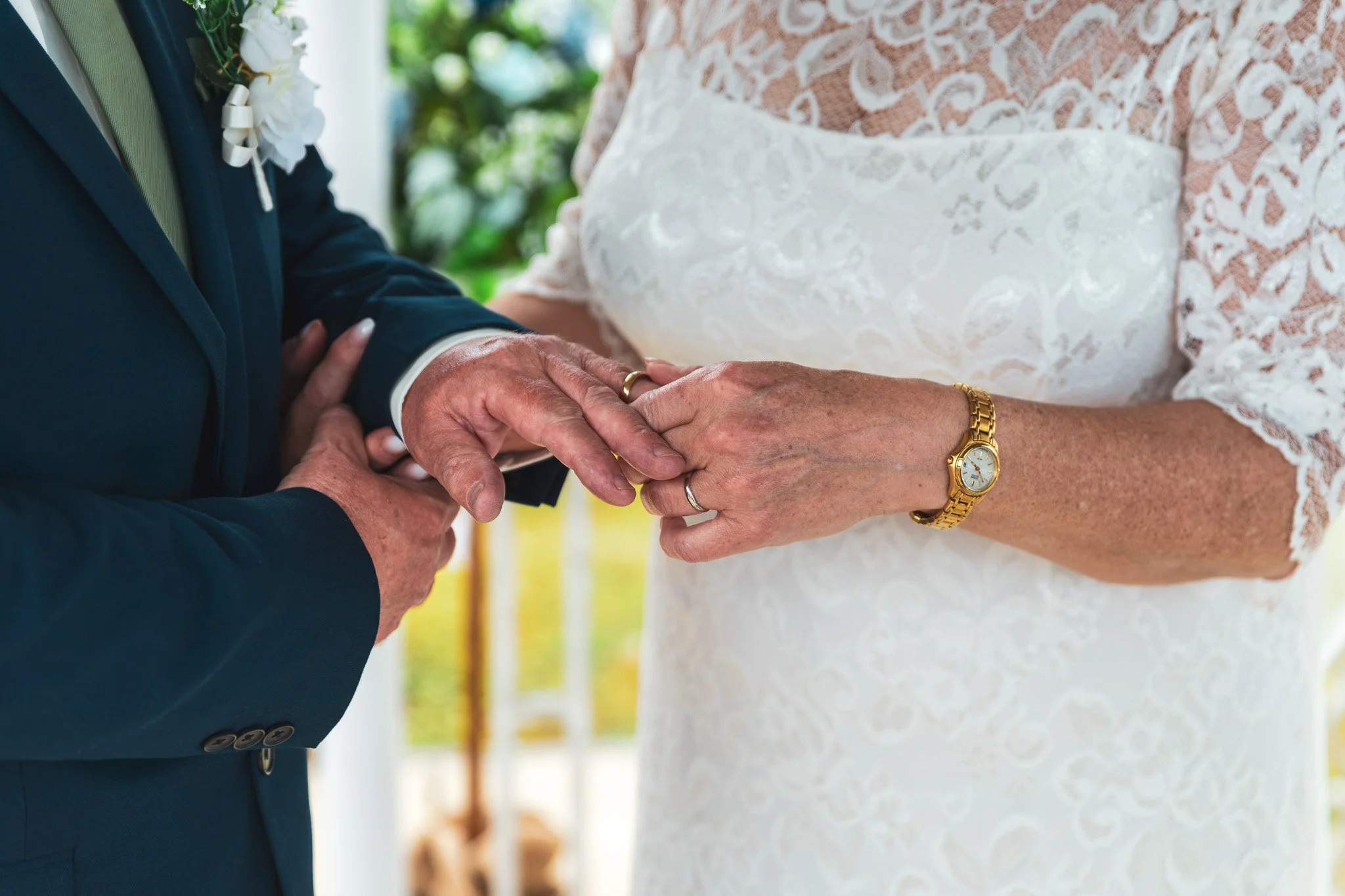 Close-up of a bride and groom holding hands during their wedding ceremony. The groom wears a dark suit, and the bride is in a white lace wedding dress with a gold watch and wedding rings visible.