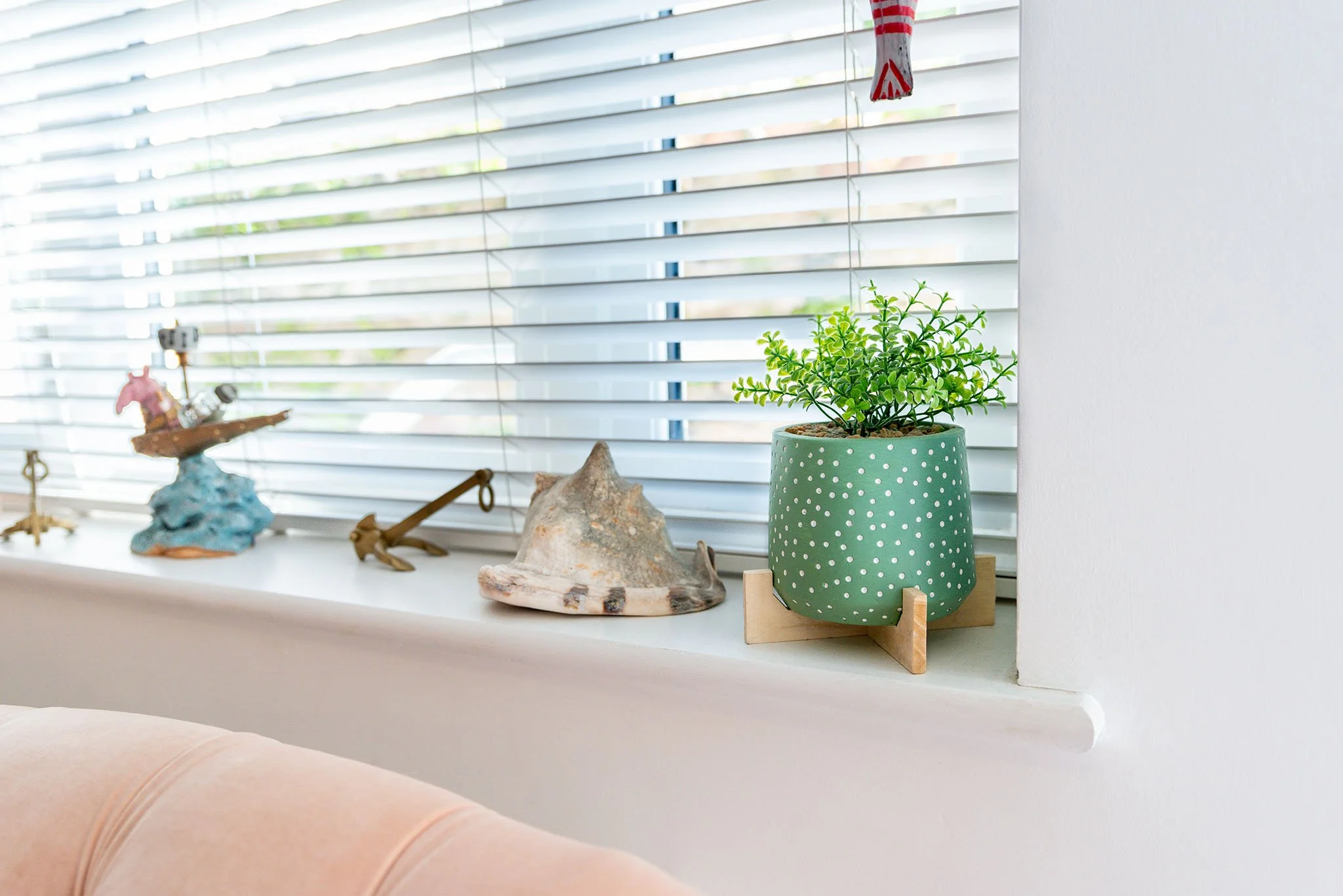 A windowsill from a Torbay holiday home with a green potted plant with small leaves in a polka dot pot, and various decorative items including a shell and figurines, in front of closed white blinds.