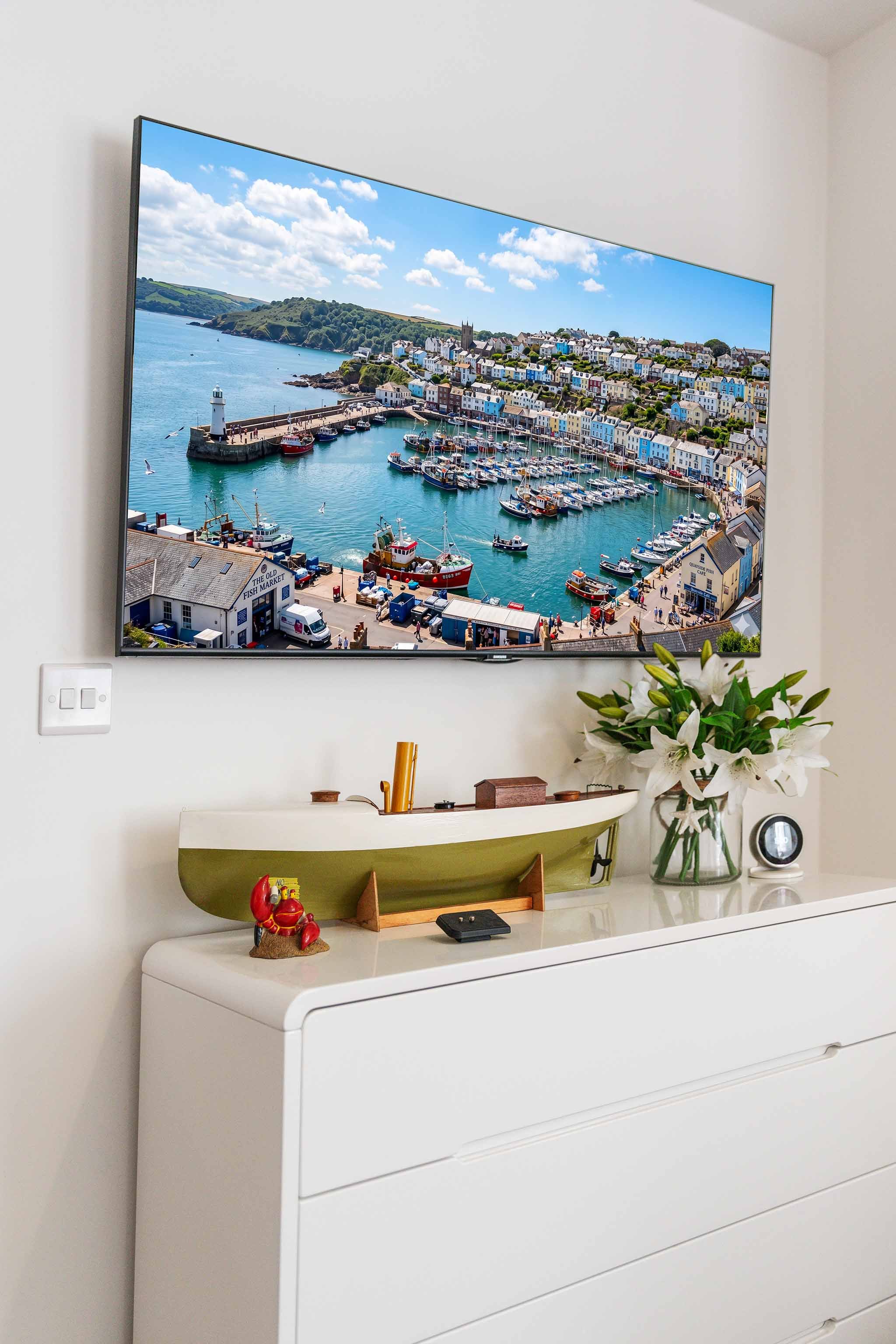 Living room with a mounted flat-screen TV showing a coastal town with boats and colourful buildings in Brixham, Torbay. Below the TV, a decorative boat model, red statue, glass vase small round speaker are arranged on a white cabinet.