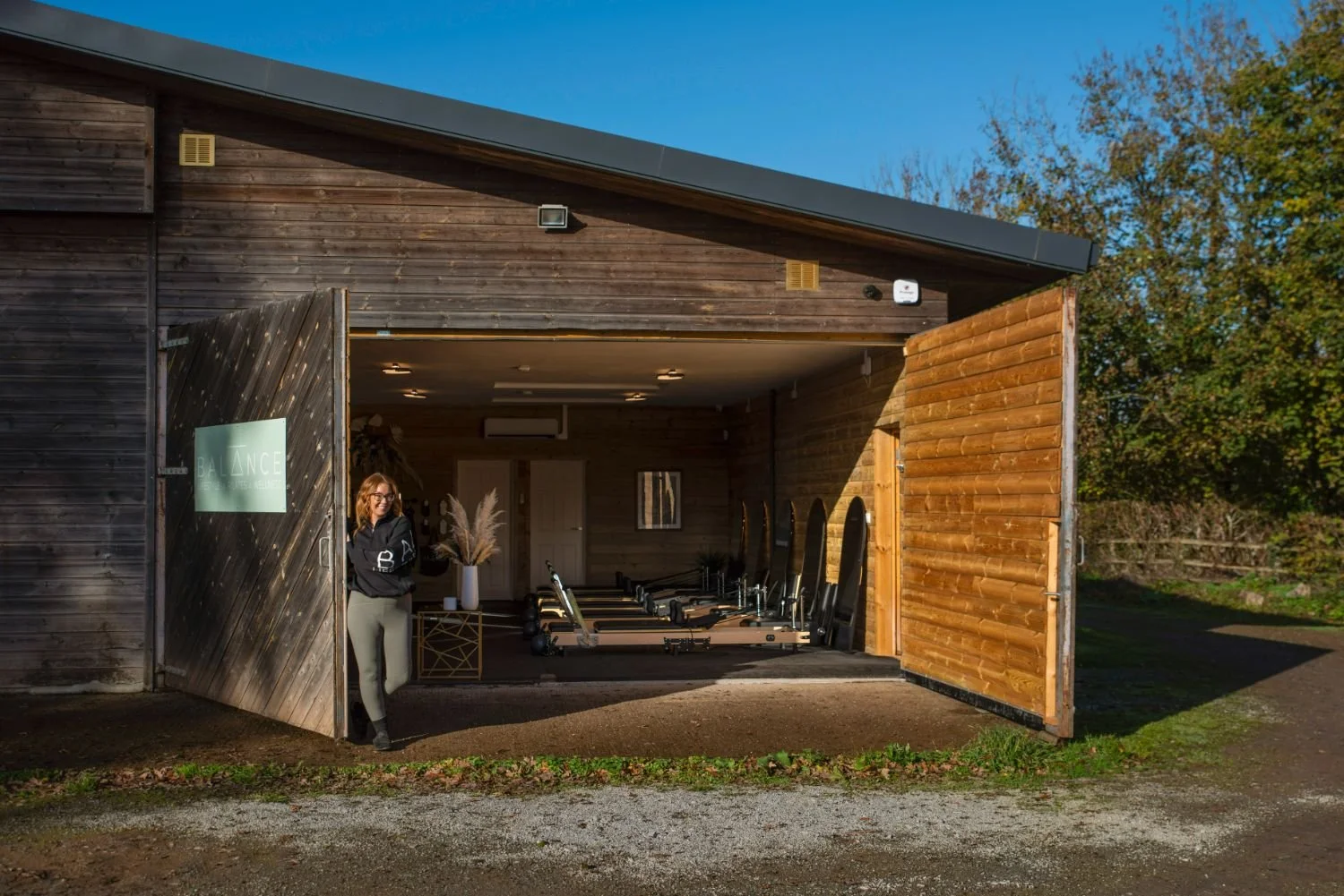 A woman standing outside an open gym with pilates reformer equipment inside, wooden walls, and a sign that reads 'BALANCE'.