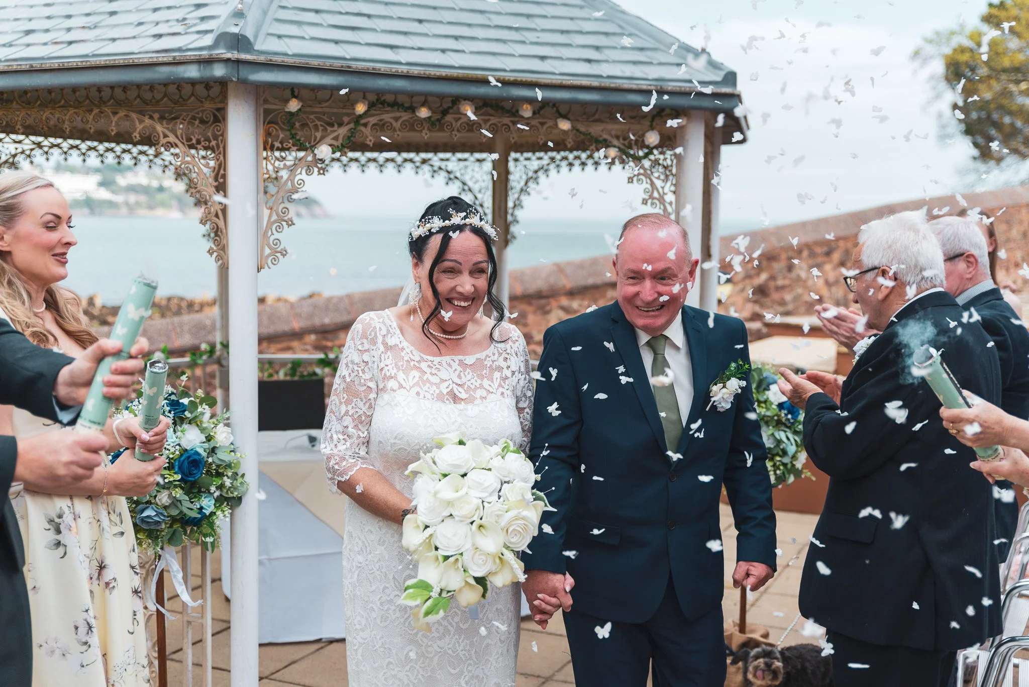 A wedding ceremony outdoors with a bride and groom holding hands under a decorated arch, surrounded by guests and white confetti, next to a body of water.