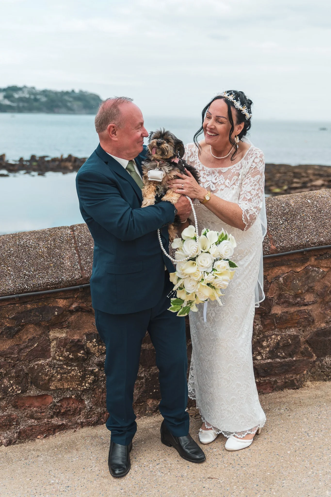 A wedding couple standing outdoors near water in Torquay at the Livermead Cliff Hotel, holding a small dog between them. 