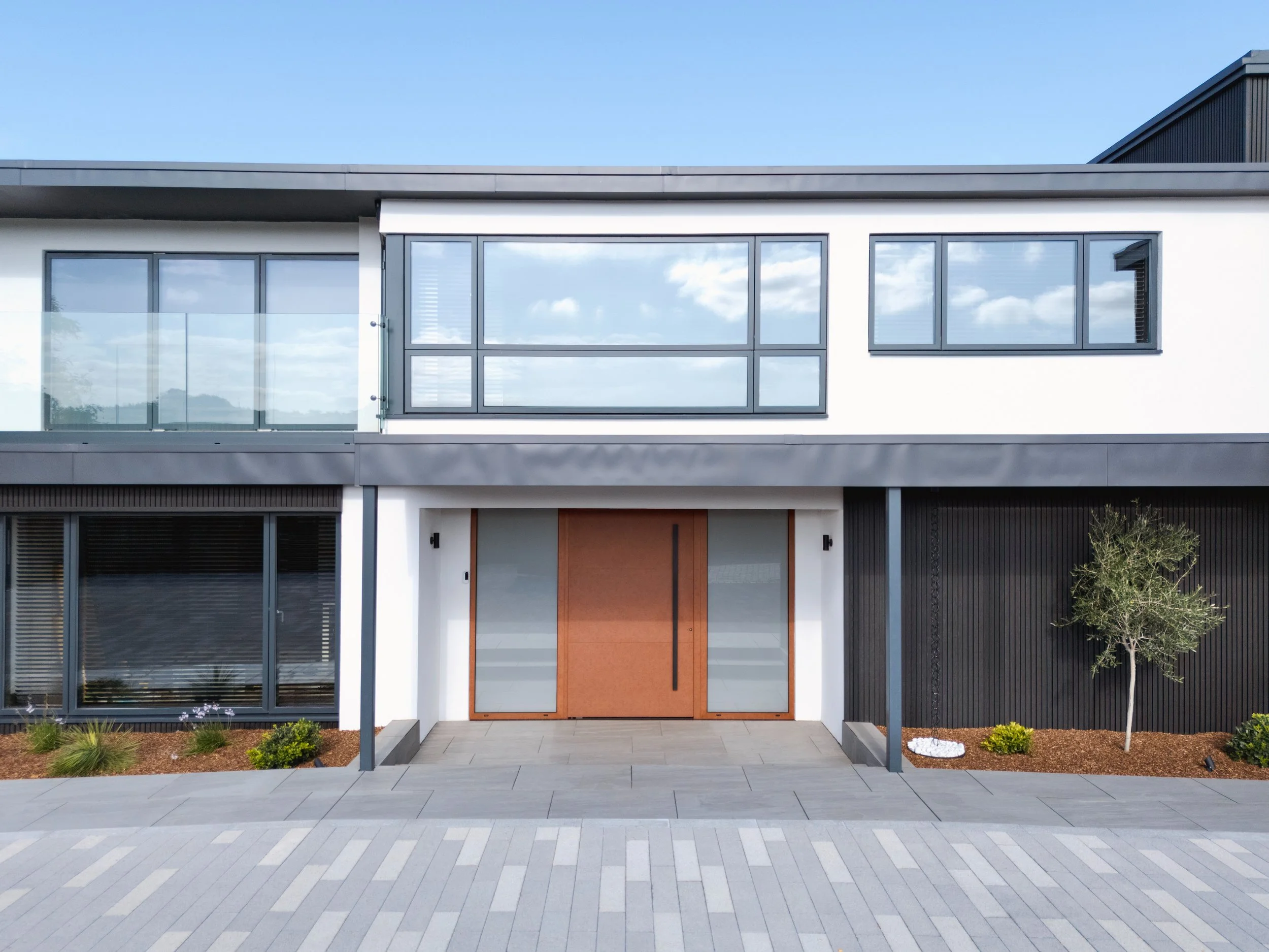 Modern two-story house with large windows, a wooden front door, and a small landscaped area with a tree and plants in front.
