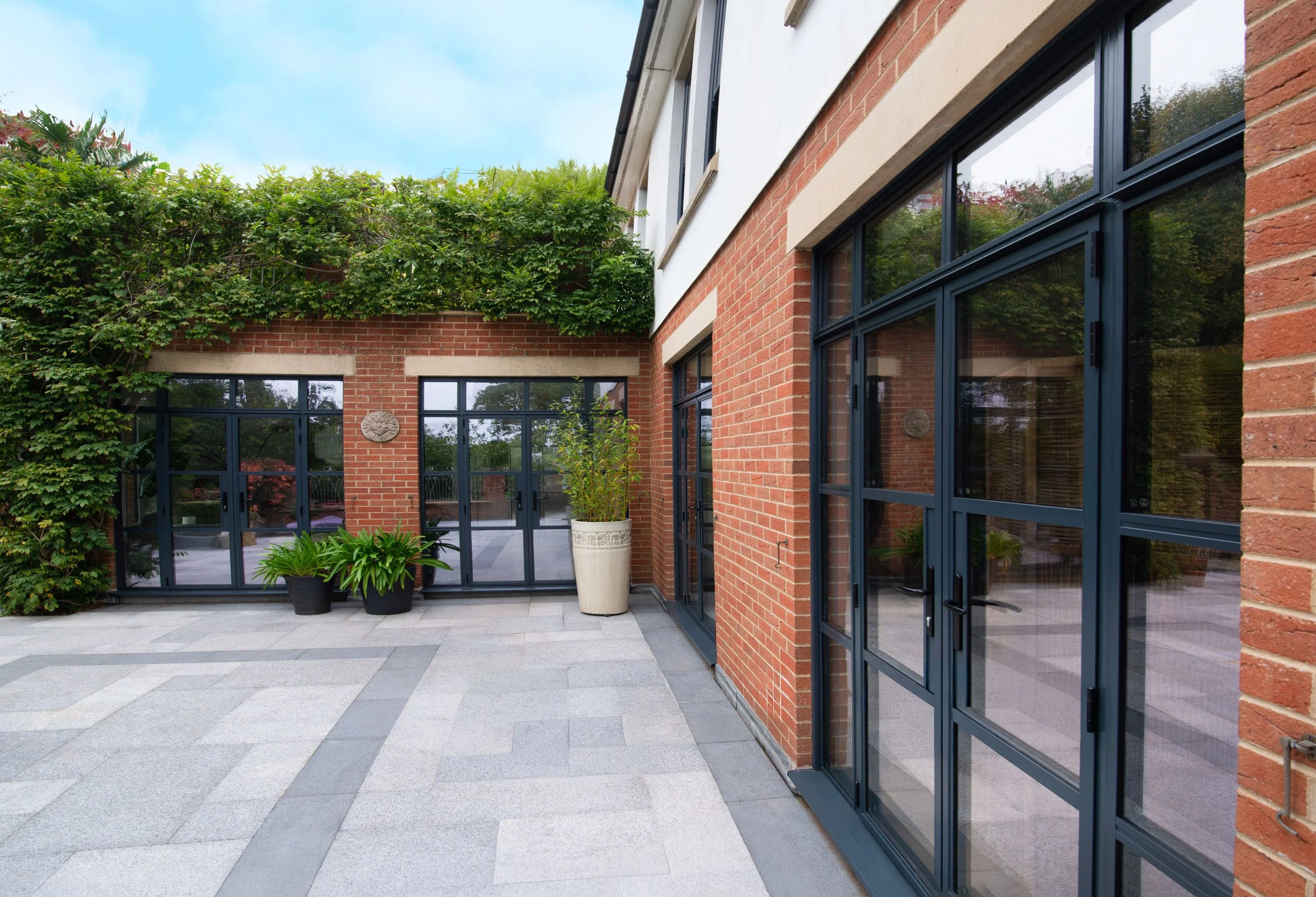 Exterior view of a modern building with red brick walls, large glass doors, and windows, potted plants, and greenery on top of the building.