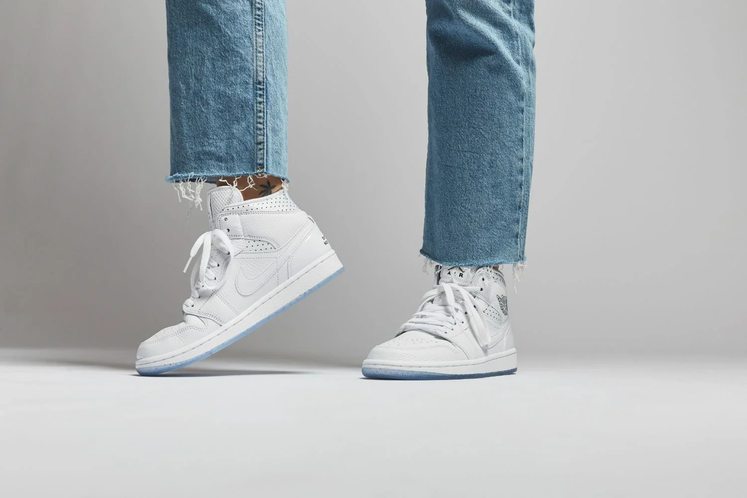 Person wearing distressed blue jeans and white Nike Air Jordan sneakers, standing on a plain white surface against a grey background.