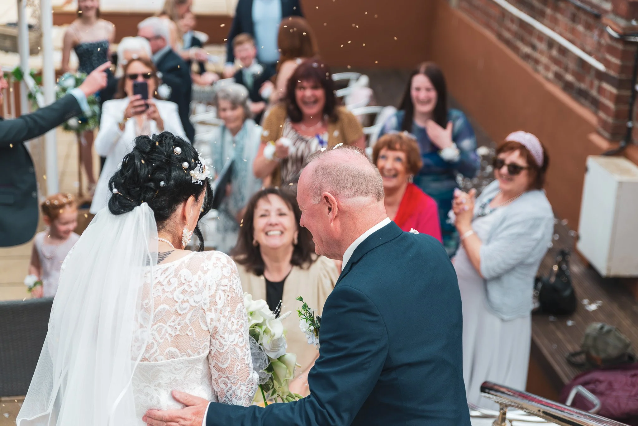 A wedding ceremony with a bride and groom exchanging vows, surrounded by guests celebrating and taking photos. The bride wears a white lace dress with a veil, and the groom wears a navy suit. Guests are smiling and dressed in casual and semi-formal a