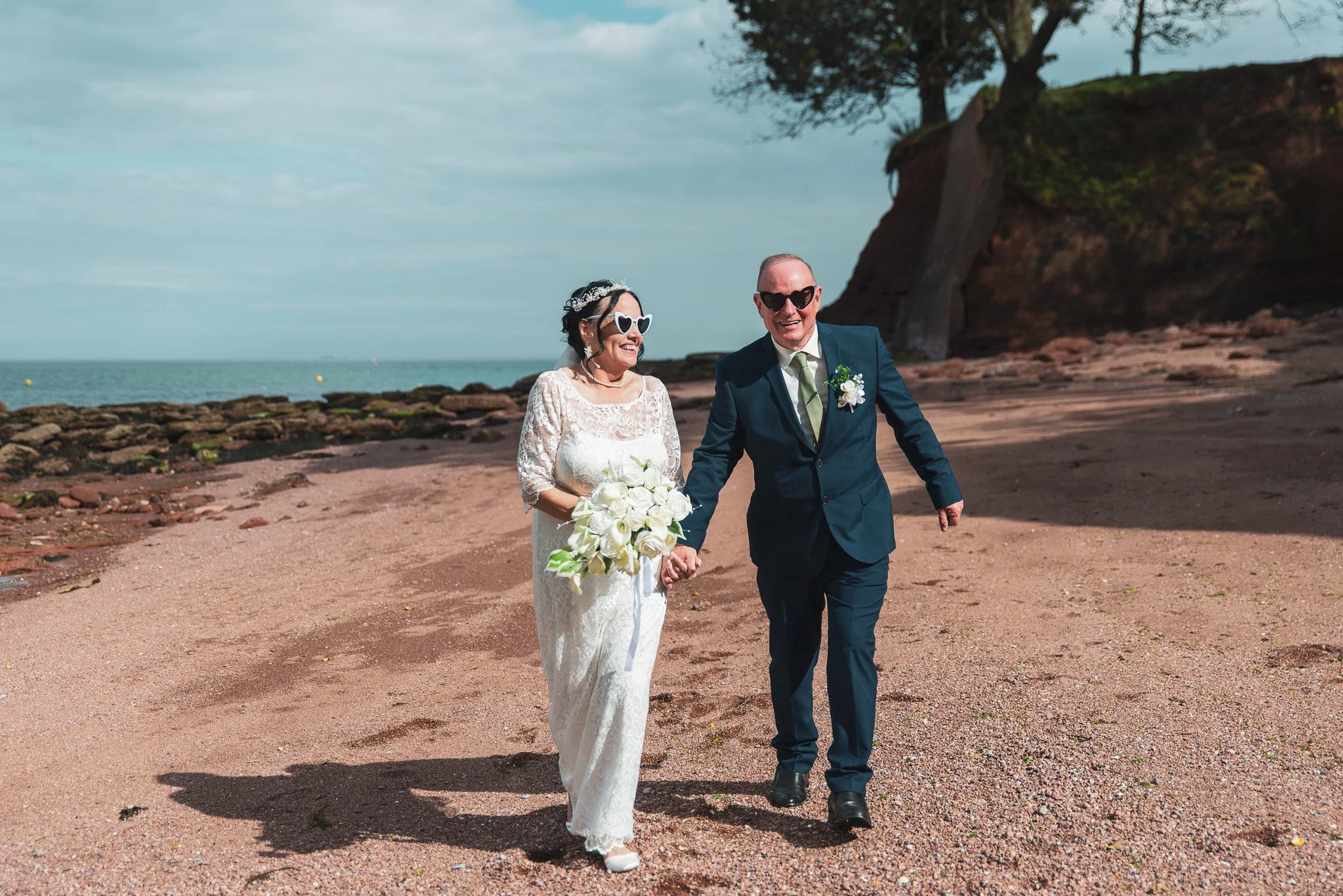 A bride and groom walking hand in hand on a beach, dressed in wedding attire, with the bride holding a bouquet of white roses, wearing heart-shaped sunglasses, and the groom wearing dark sunglasses, with a large tree and cliff in the background.