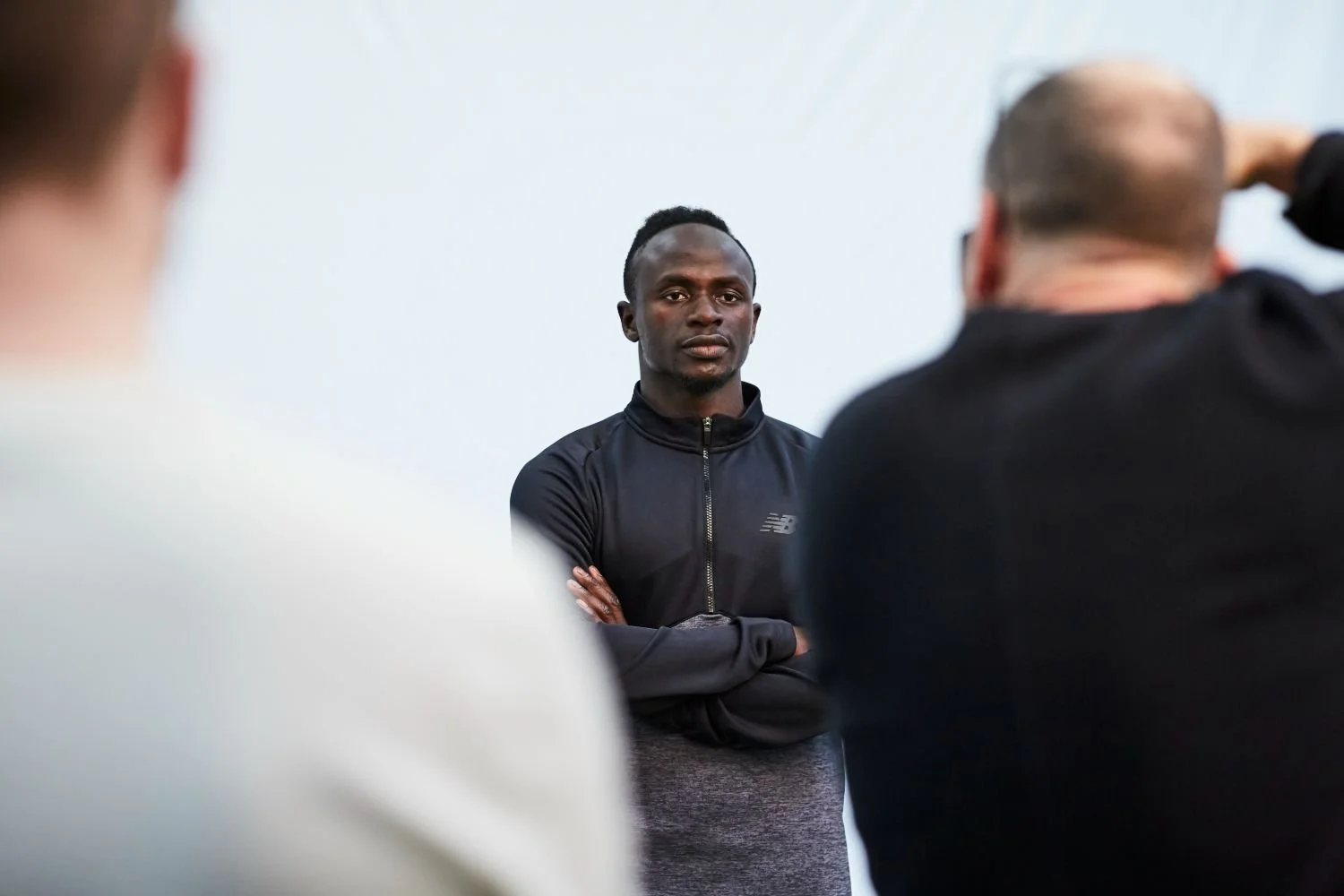 A young man wearing a black athletic jacket with folded arms listening intently as another person gestures in front of him during an indoor gathering.