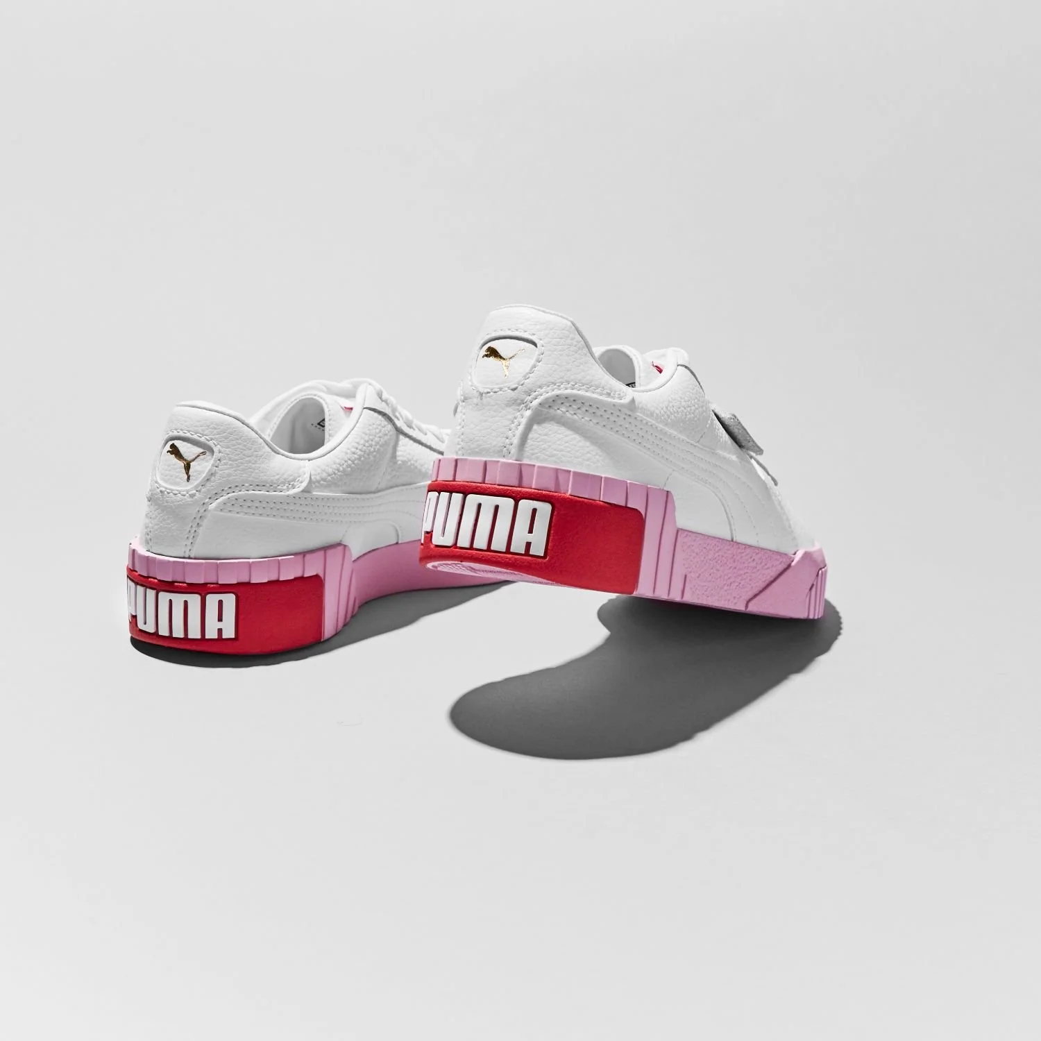 White Puma sneakers with pink and red accents, featuring the Puma logo on the heel and side, placed on a gray surface with shadows.
