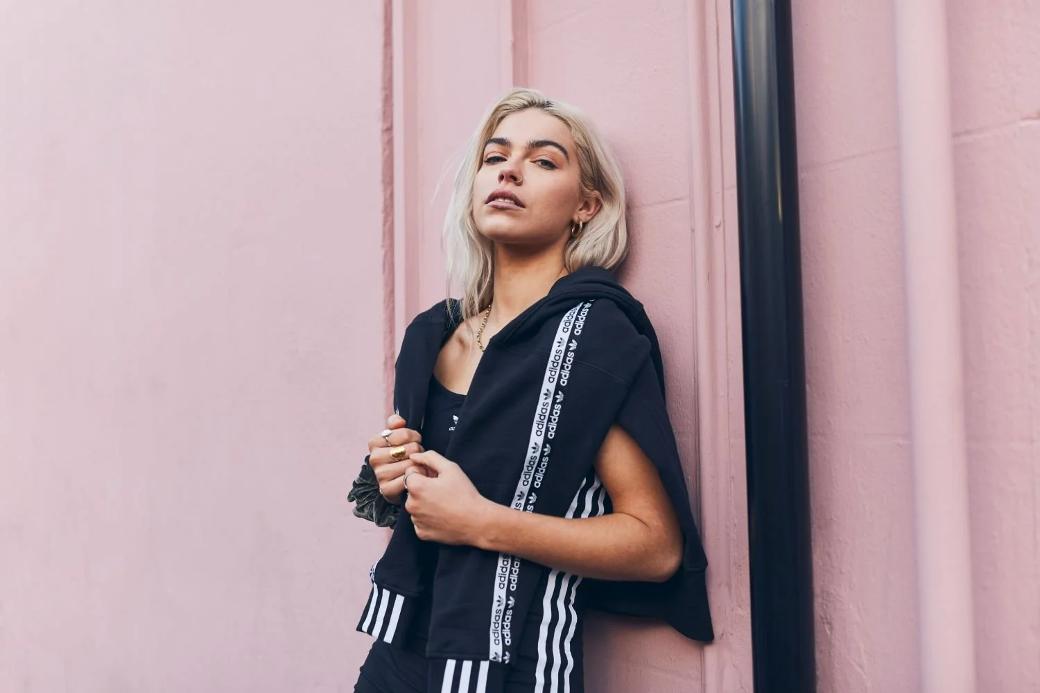 Young woman with platinum blonde hair leaning against a pink wall, wearing a black Adidas jacket with white logo stripes.