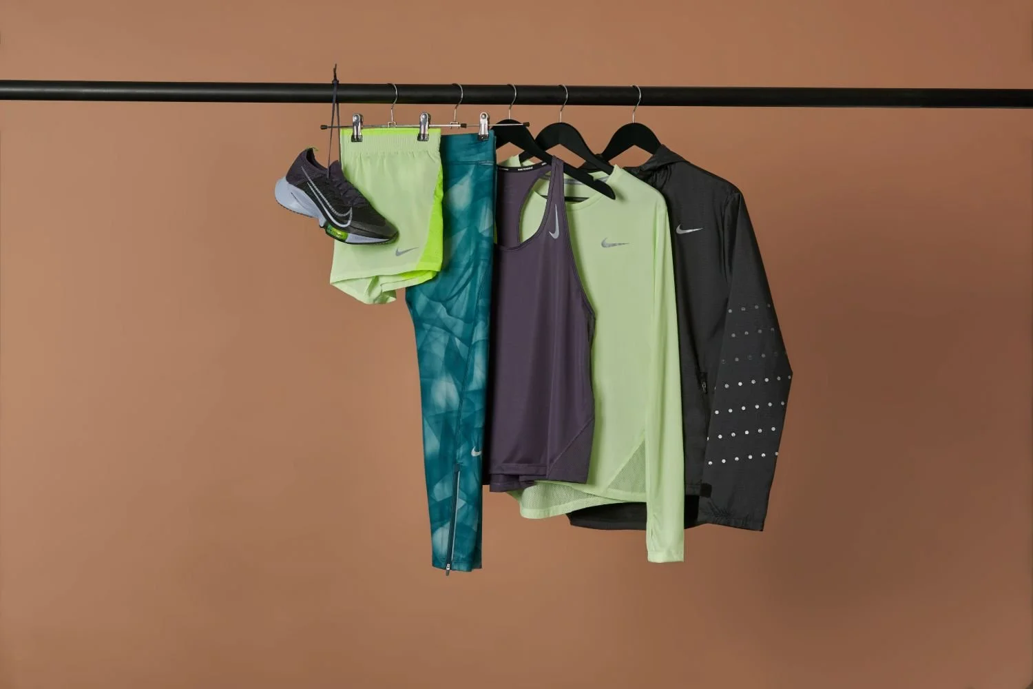 Clothing and running shoes hanging on a black rack against a plain brown background. Items include two pairs of shorts, two long sleeve shirts, a tank top, a jacket, and a pair of black running shoes.