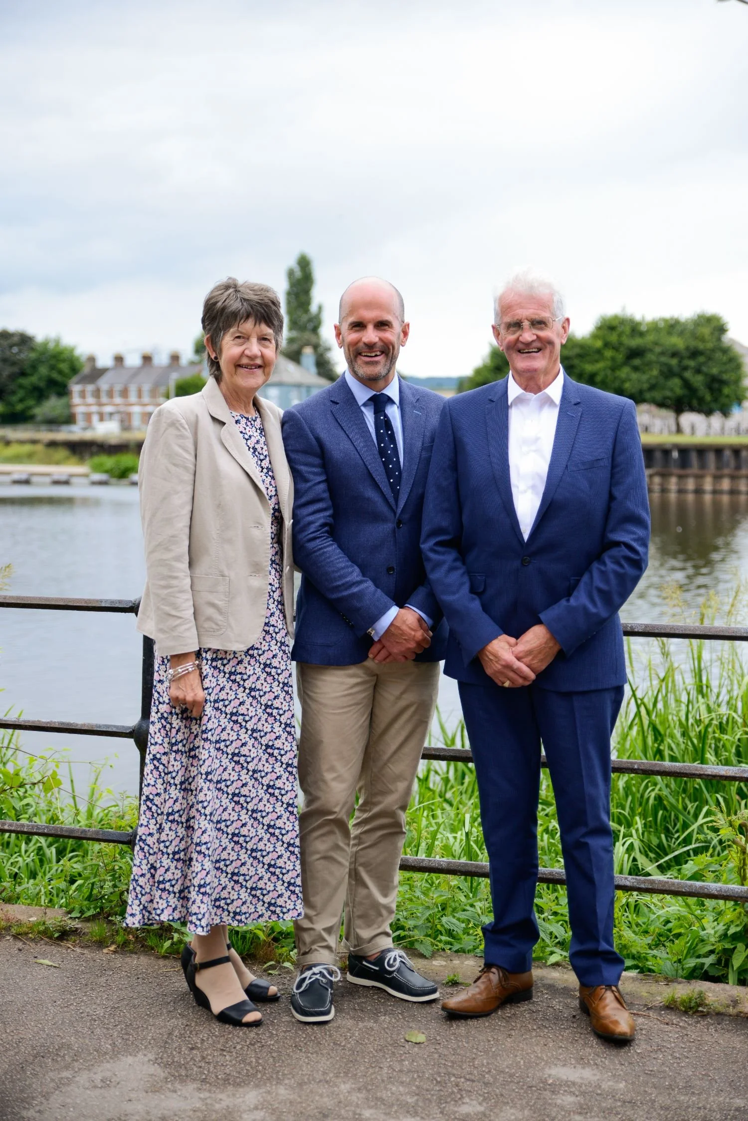 Three people, two men and one woman, dressed in formal attire, standing outdoors near a river with houses and trees in the background, smiling at the camera.