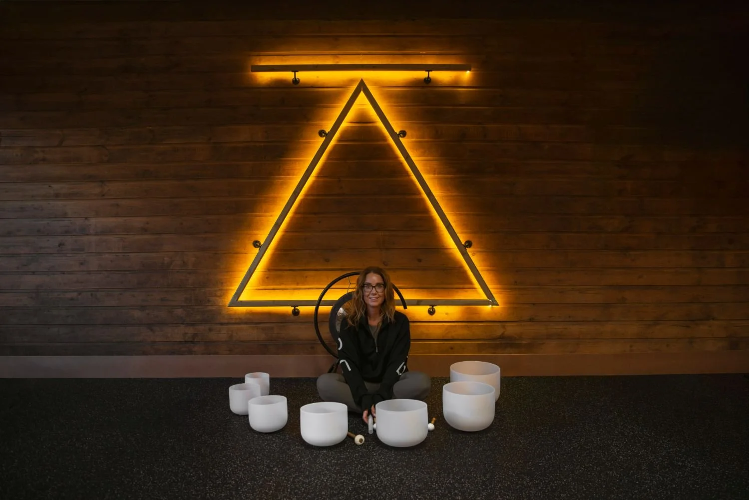 A woman sitting on the floor in front of a large illuminated triangle on a wooden wall, surrounded by white singing bowls, with a dark carpeted floor.