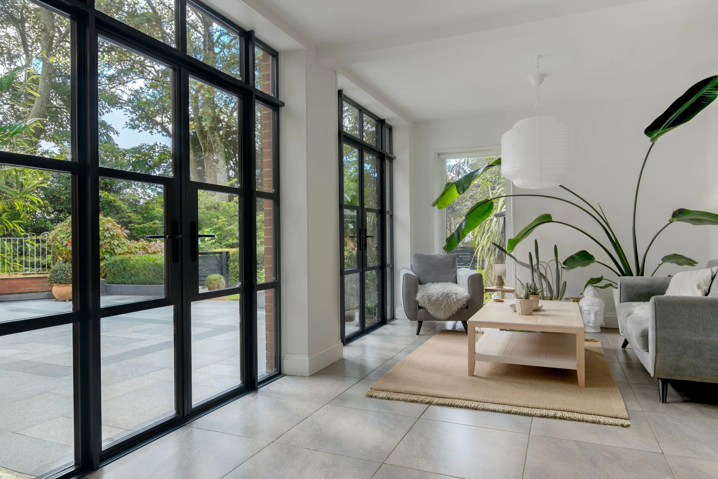 Interior view of a bright living room with large windows, a gray armchair with a furry throw, a light wood coffee table, a beige rug, and several potted plants, including a large one with big green leaves.
