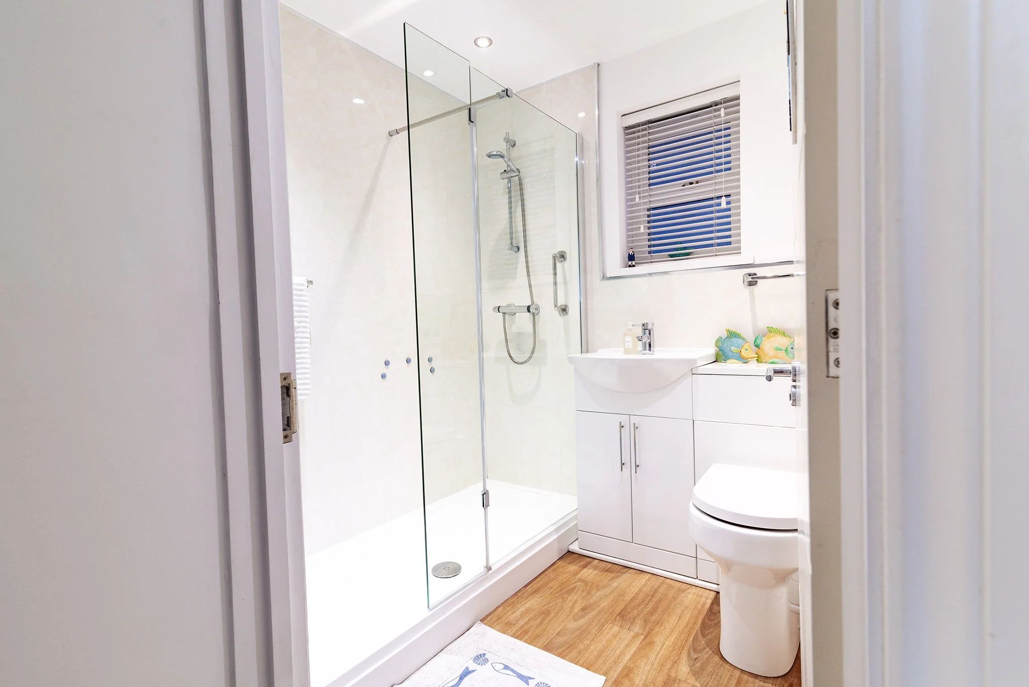 A modern bathroom in Torbay based holiday home with a glass shower enclosure, a white toilet, a small white vanity with a sink, a window with blinds, and some colourful fish decorations on the counter.