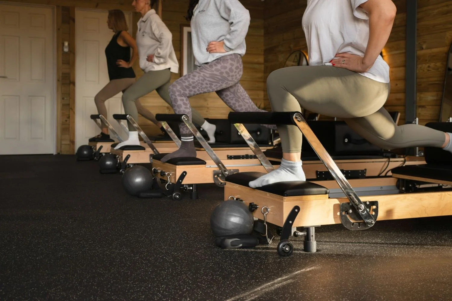 Four women in workout clothing doing high knees on wooden exercise steps in a gym with black flooring and wood-paneled walls.