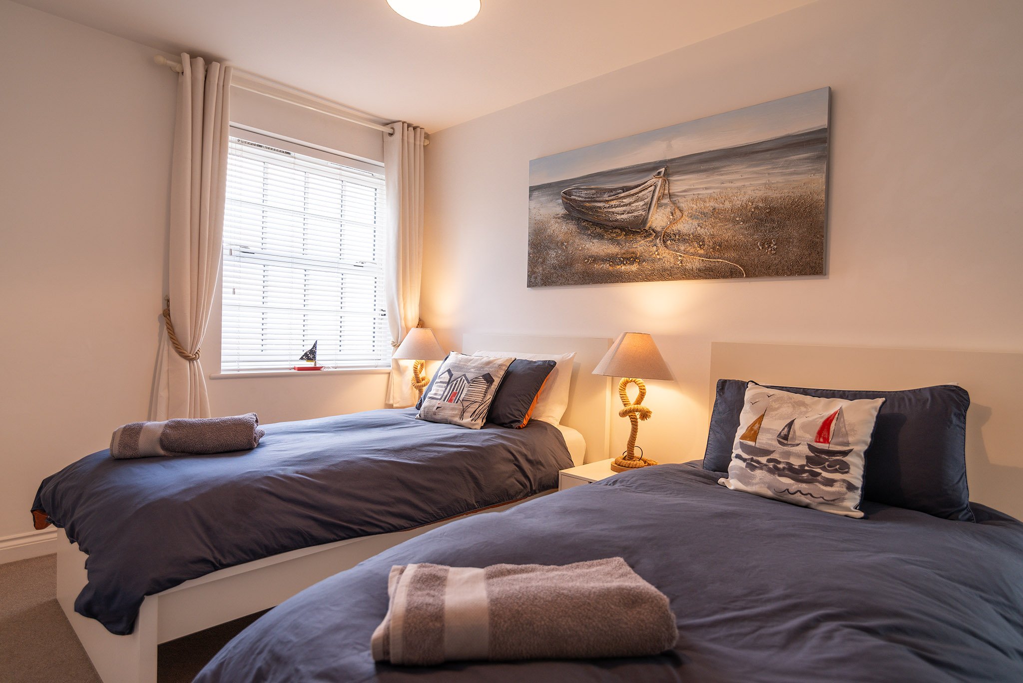 Bedroom of a holiday let in Brixham, Torbay, with two beds, navy bedding, nautical-themed pillows, and a wall-mounted painting of a boat on the beach, with natural light coming through white blinds and cream curtains.