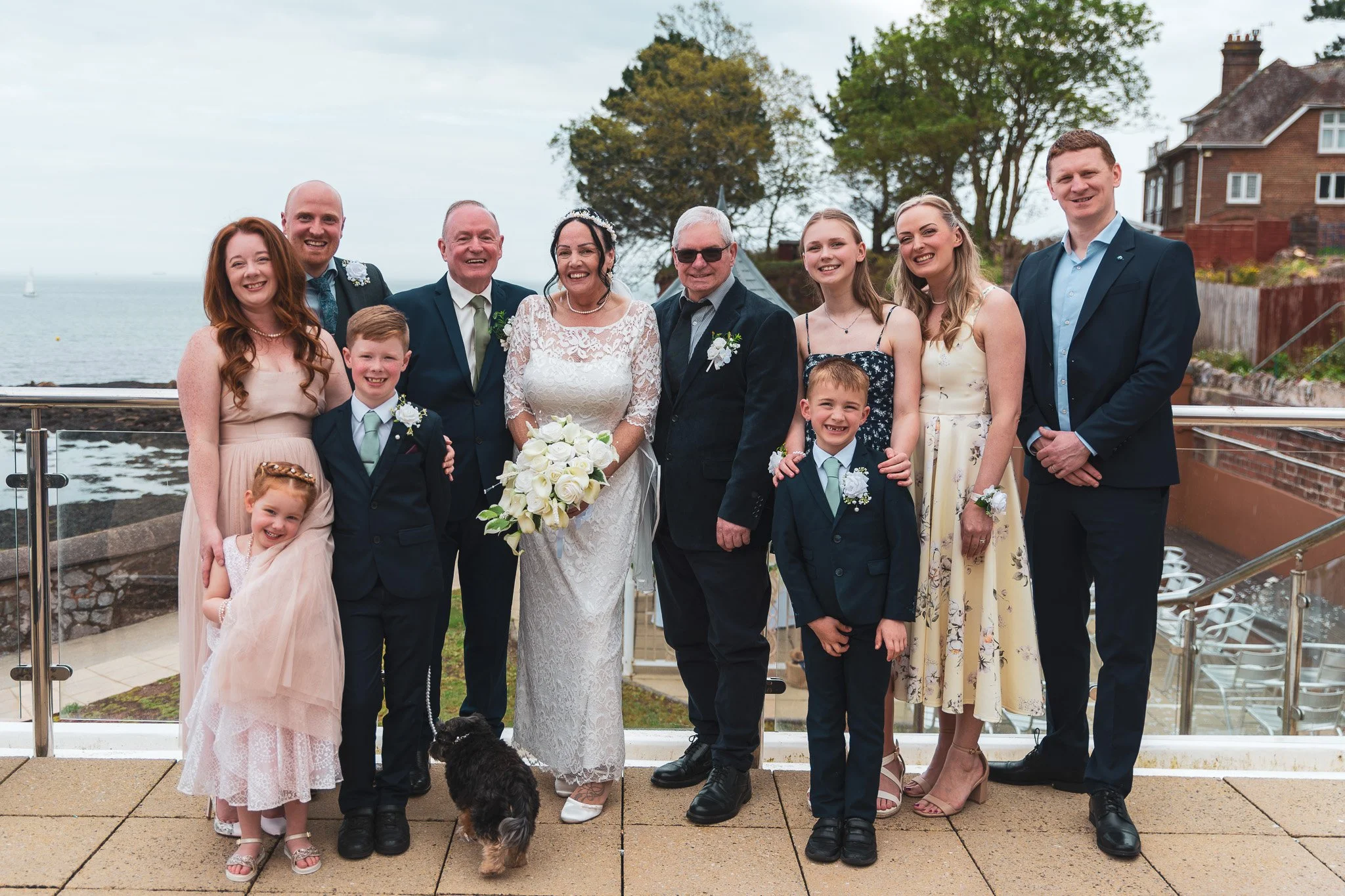 A group of people dressed in formal attire at a wedding by the water in Torquay at the Livermead Cliff Hotel. They are smiling and standing outdoors with a view of trees, houses, and boats in the background.
