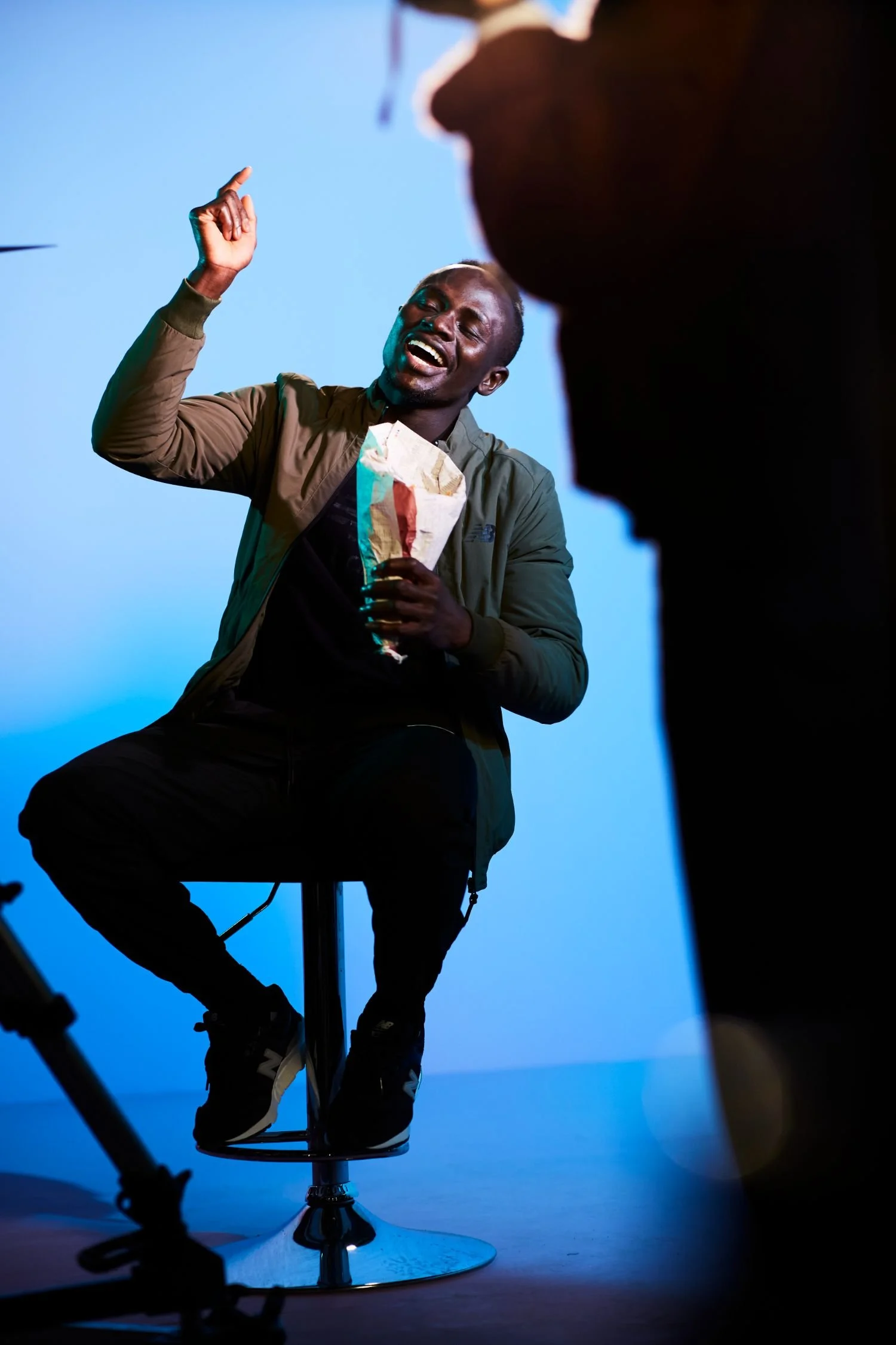 A man sitting on a stool, laughing and gesturing with his hand, holding a paper bag, in a studio with a blue background.