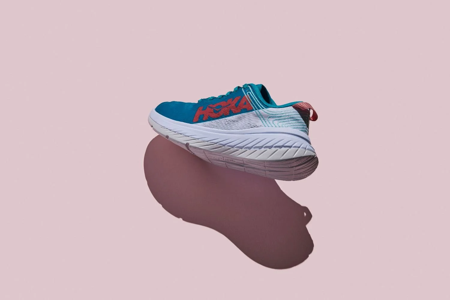 A running shoe with a blue upper, red logo, and white sole, placed on a pink background with a shadow beneath it.