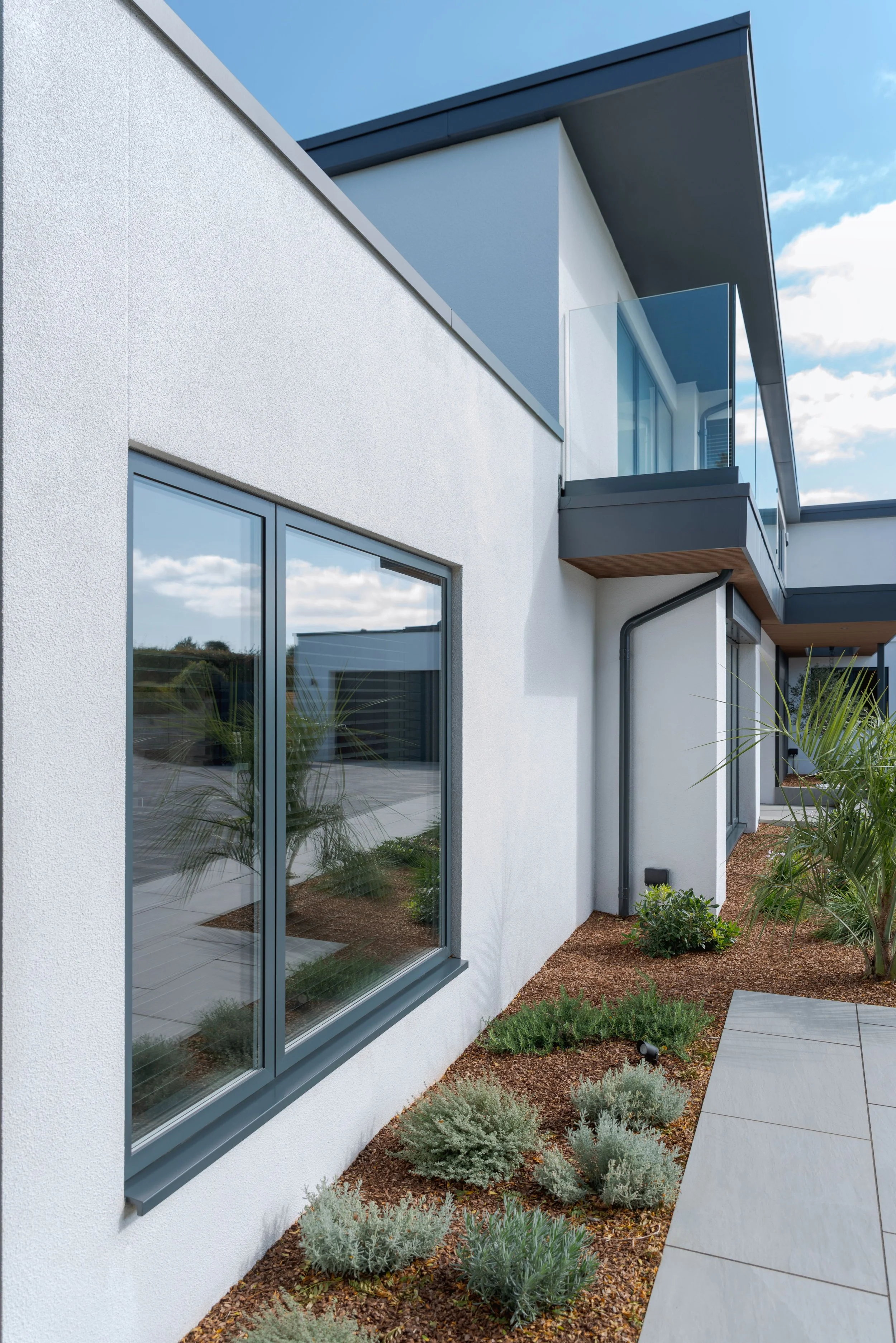 Modern residential building with white exterior walls, large glass windows, and a balcony with glass railing. Landscaping includes small shrubs, palm plants, and a tiled walkway.