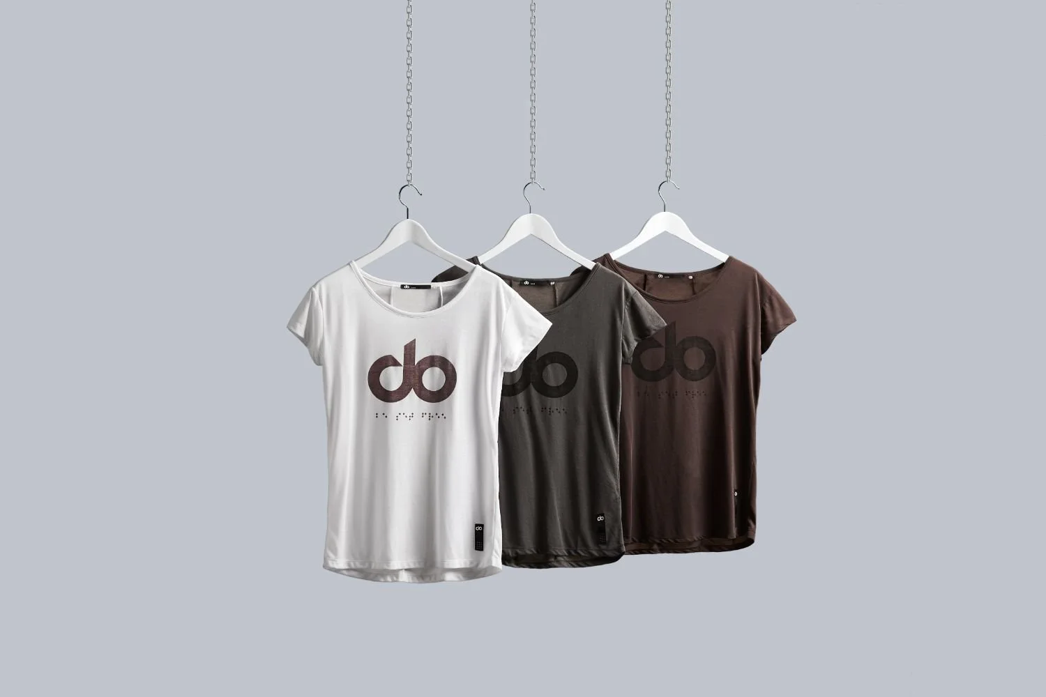 Three T-shirts in white, gray, and brown hanging on white hangers against a light gray background. All display a large black 'do' and smaller Braille underneath.