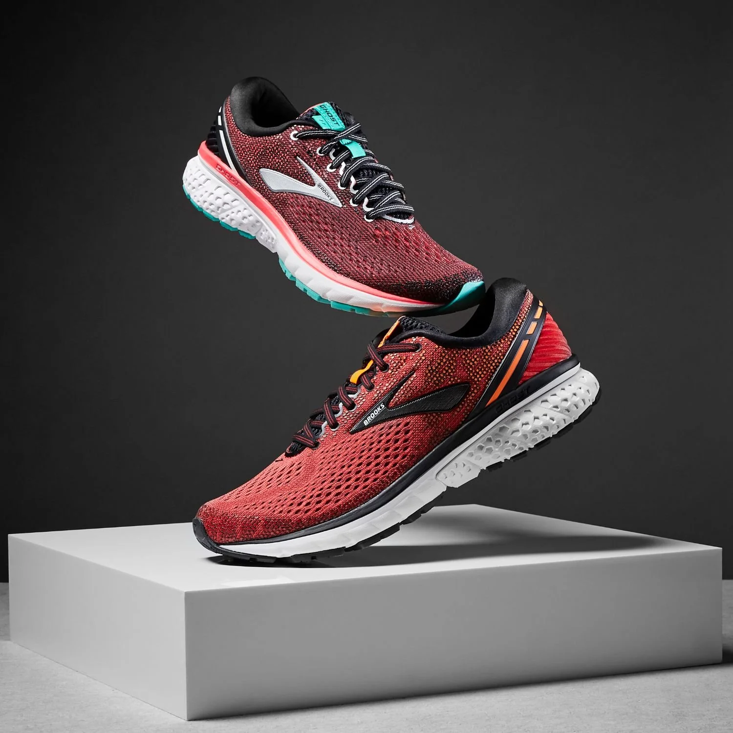 Two red running shoes with black laces and white soles are displayed on a white pedestal against a dark background.