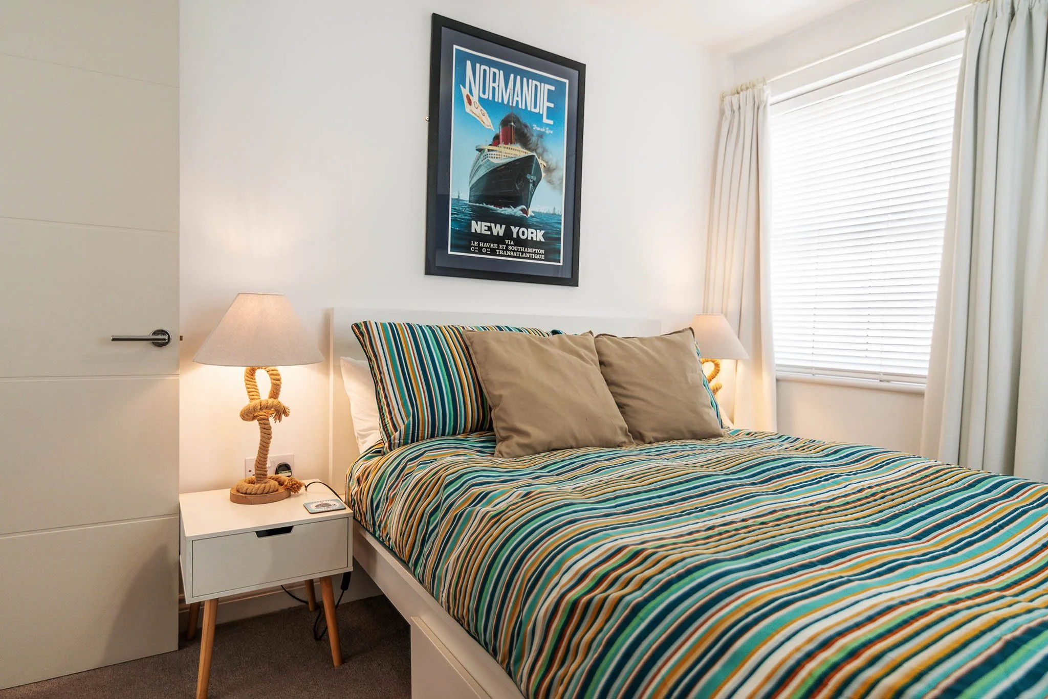 Holiday let in Brixham with a bedroom with a bed that has a striped bedspread and pillows, a white nightstand with a lamp, a wall-mounted framed poster of a ship titled 'Normandie' above the bed, and closed white blinds on a window with curtains.