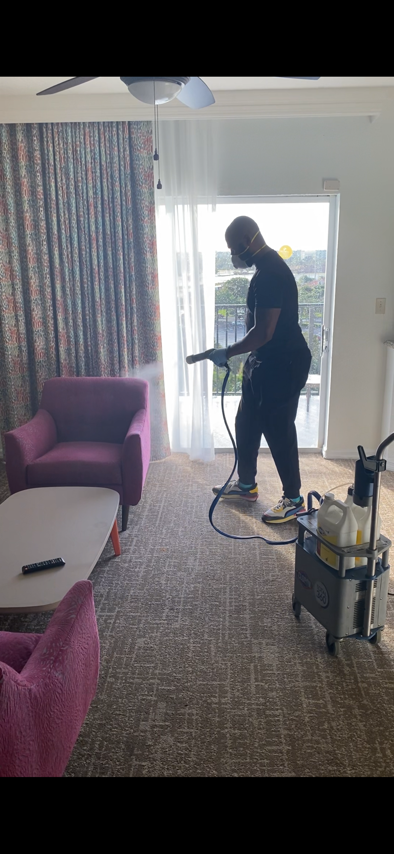 A person wearing a face mask and gloves is disinfecting a hotel room with a steam cleaner near a balcony door. The room has pink chairs, a white coffee table, and patterned curtains.