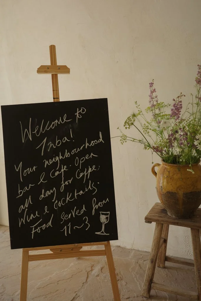 Blackboard sign welcoming visitors to a neighborhood bar and café, with hours from 11 a.m. to 3 p.m., and a small drawing of a wine glass below the text.