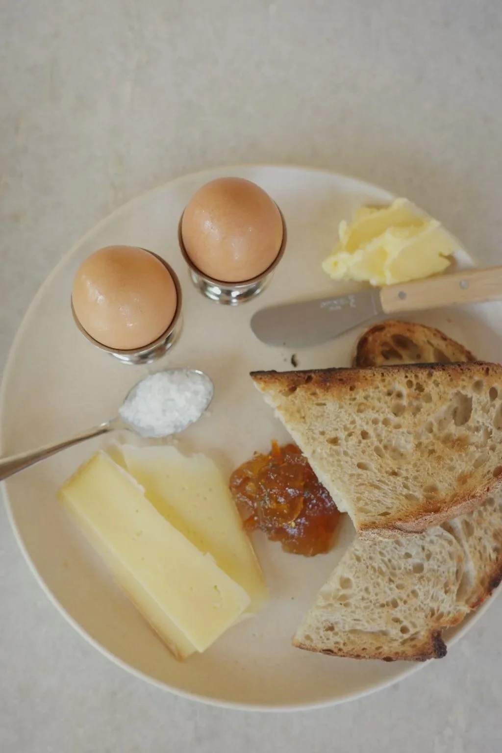 A breakfast plate with two eggs in egg cups, slices of bread, butter, jam, a piece of cheese, and a teaspoon of salt.