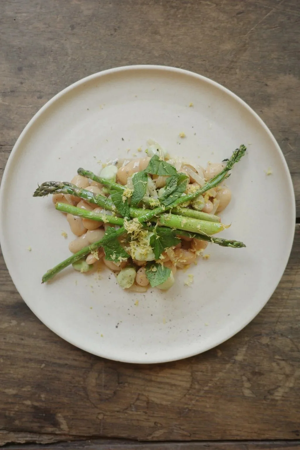 A white plate with cooked green asparagus, leafy herbs, chickpeas, and grated lemon zest on a wooden surface.