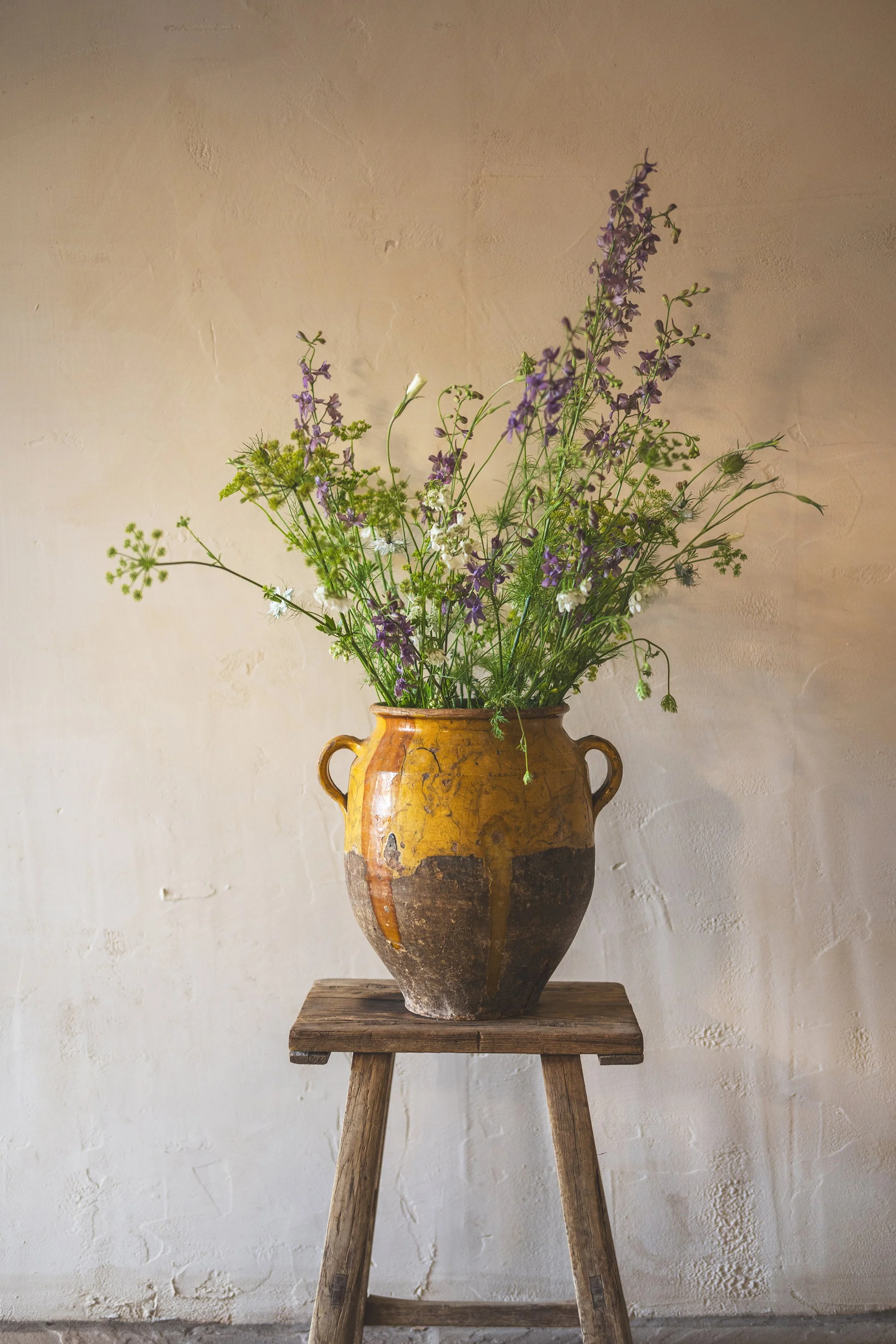 A large, rustic ceramic vase with two small handles, filled with a wildflower arrangement featuring purple, white, and green flowers, sits on a wooden stool against a textured off-white wall.