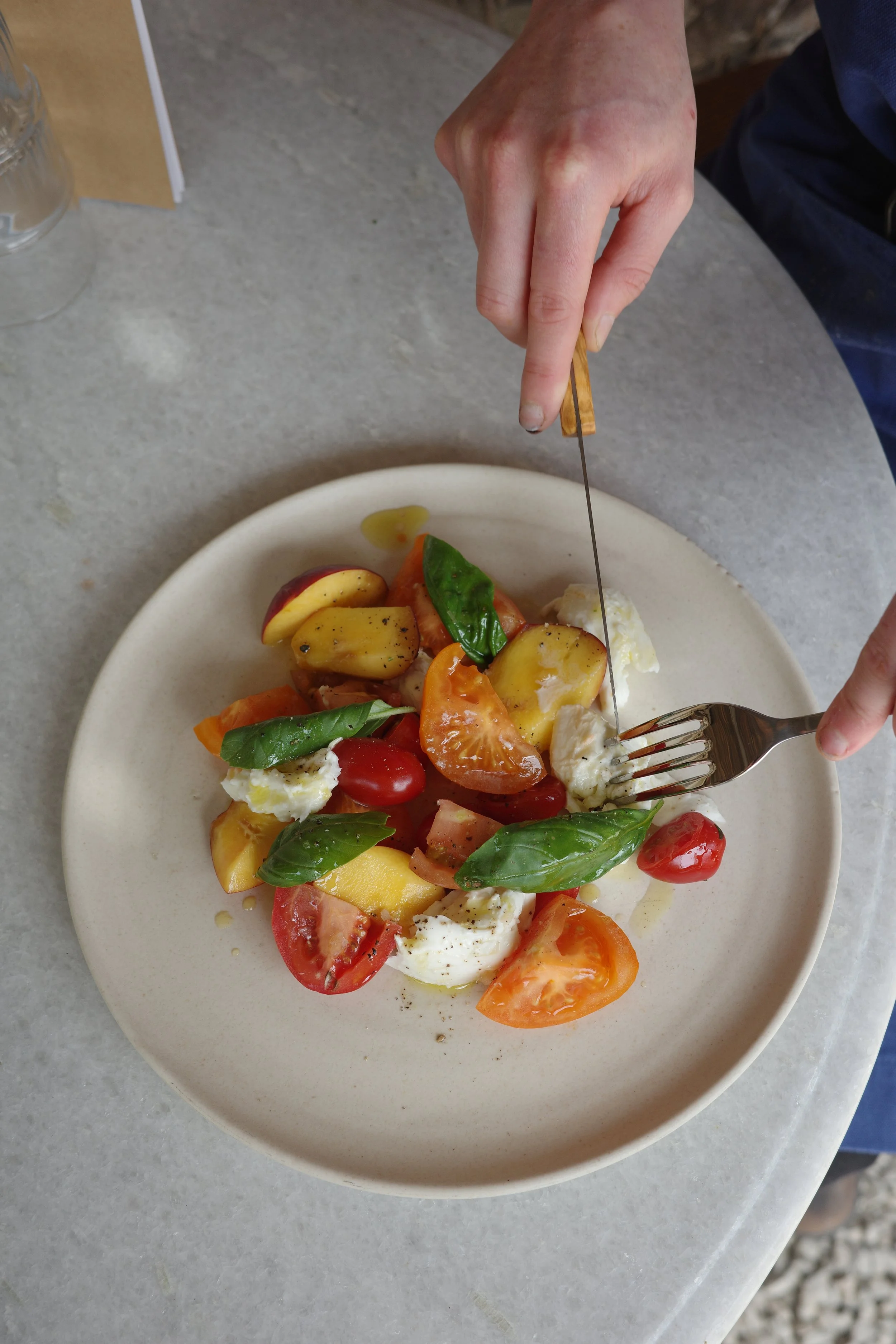 A plate of fresh tomato and mozzarella salad garnished with basil leaves on a white plate, with a person slicing the salad with a fork and knife.