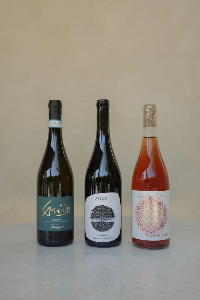 Three bottles of wine on a light-colored surface against a plain beige background, with the middle bottle slightly taller.