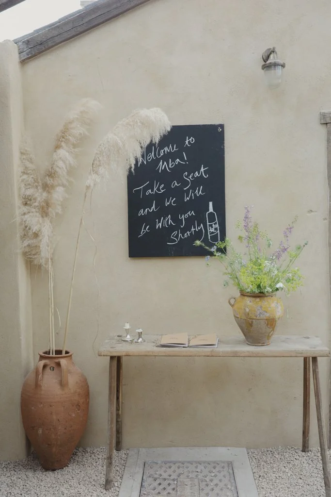 A wall with a chalkboard sign saying "Welcome to Nabal! Take a seat and we will be with you shortly" with a drawing of a bottle. Decor includes a large clay vase with pampas grass on the left and a ceramic vase with flowers on a wooden table.