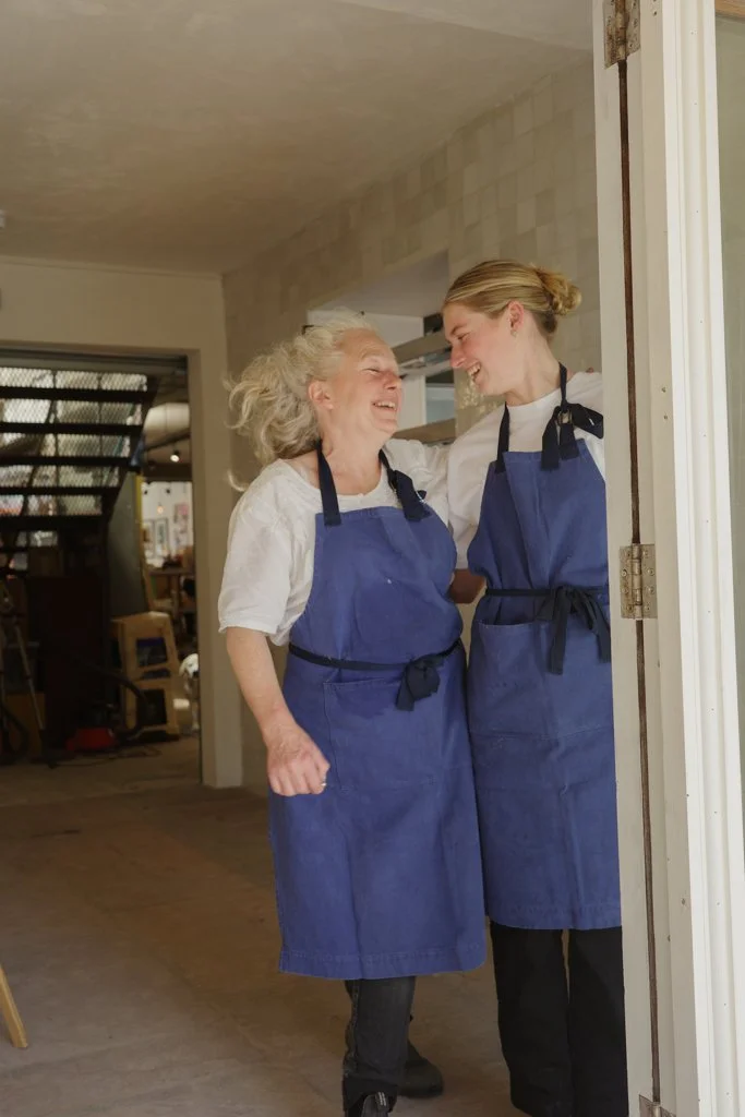 Two women, likely a mother and daughter, wearing blue aprons are happily embracing and smiling at each other in a cozy indoor kitchen or restaurant setting.