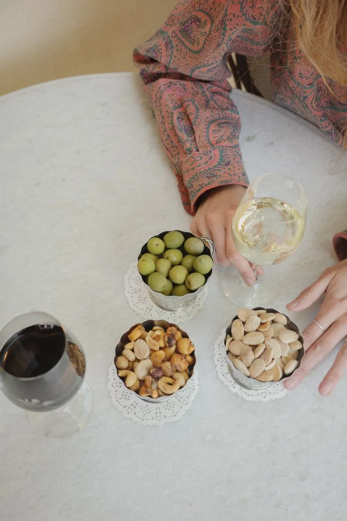Person with long blonde hair wearing a patterned jacket holding a glass of white wine, surrounded by three bowls of snacks and a glass of red wine on a white table.