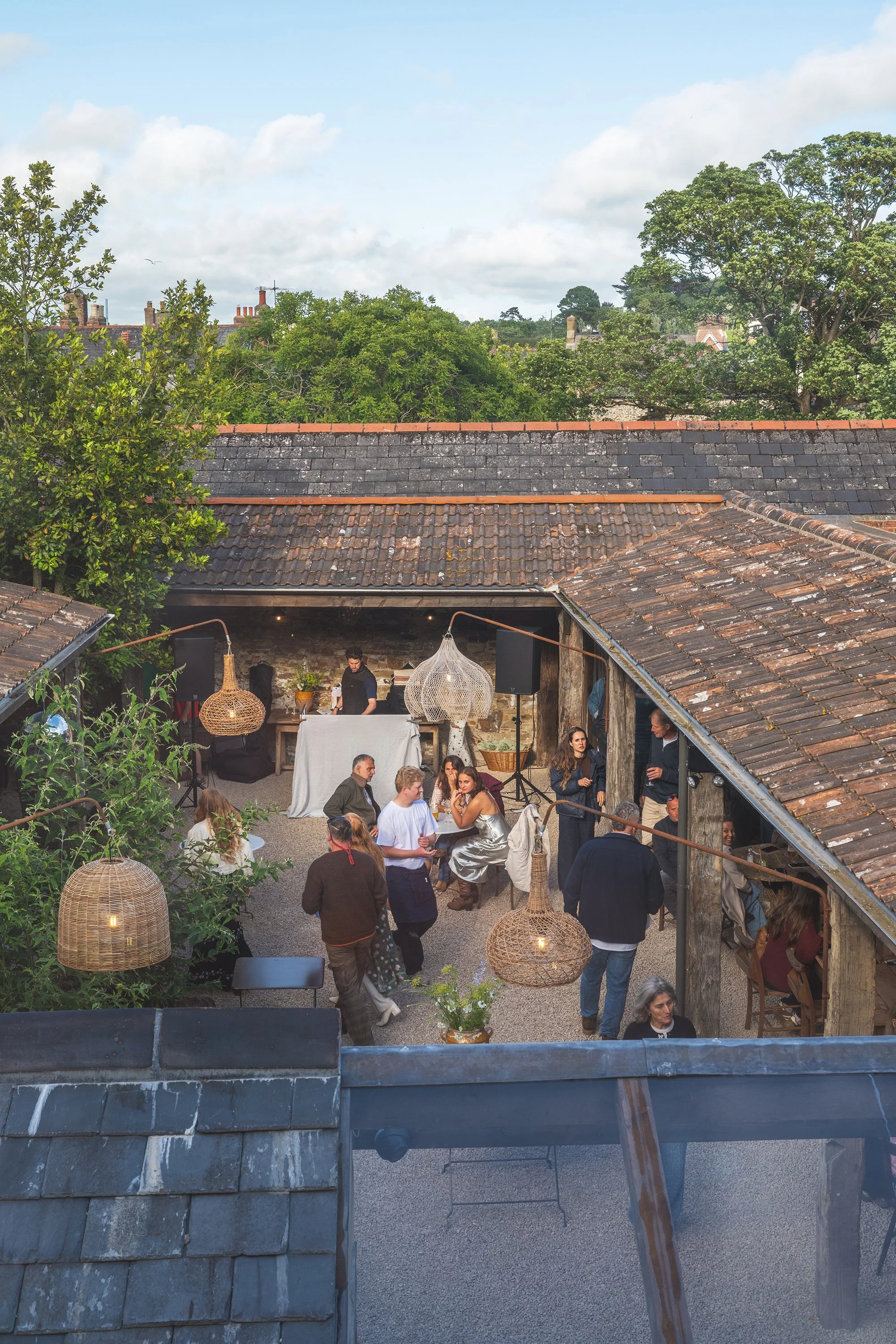 A lively outdoor gathering with people socializing, sitting, and standing in an open courtyard surrounded by rustic brick buildings and rooftop tiles, decorated with large woven pendant lamps, lush green trees, and a sky with scattered clouds.